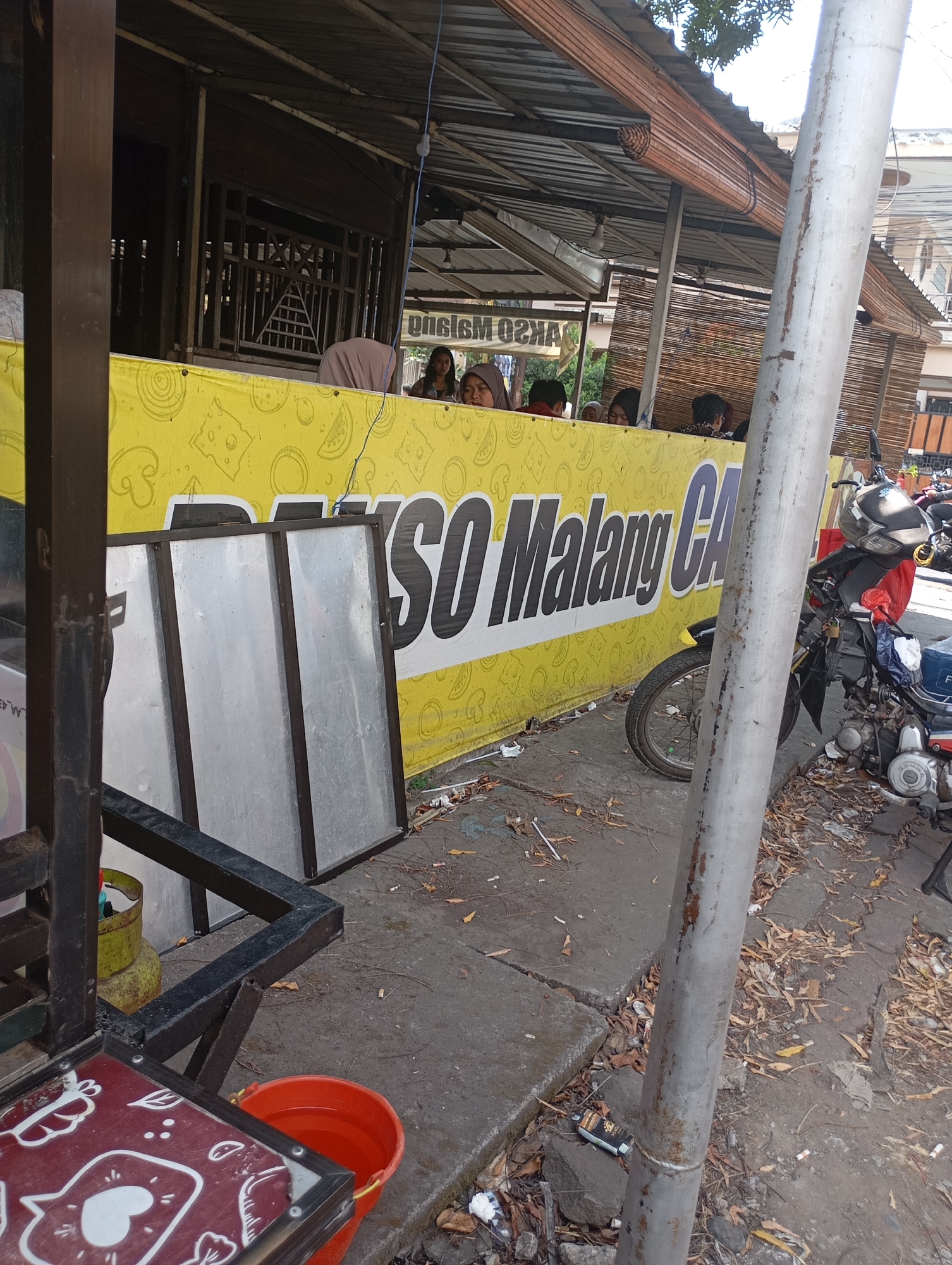 The image shows an outdoor food stall in Malang, Indonesia, likely a "bakso" (meatball soup) stall, suggested by the partial text on a large yellow banner which reads "SO Malang CA". The stall features a corrugated metal roof supported by wooden beams, with bamboo blinds on the side. Several people are seated under the shelter, some appearing to be customers. In the foreground, a metal frame structure with a red-topped table is visible, and to its left, a green gas cylinder stands on the ground. To the right of the banner, a motorcycle is parked. The ground is paved with concrete and covered with fallen leaves and debris. The overall scene depicts a typical street-side food vendor in a warm climate.