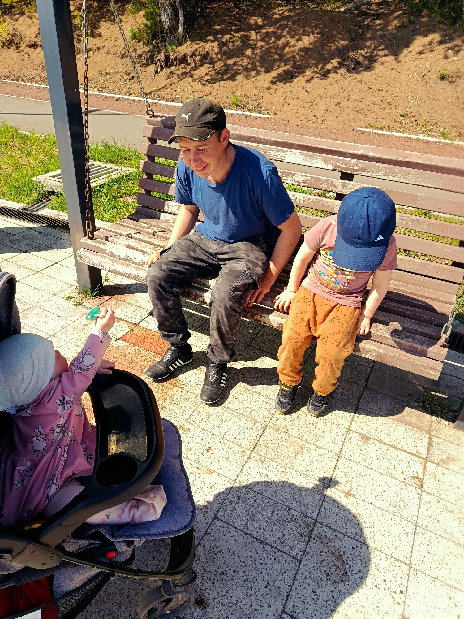 The image depicts an outdoor scene in what appears to be a park in Chaikovsky, Russia, on a sunny day.
A man wearing a blue T-shirt, camouflage pants, and a black baseball cap sits on a wooden bench. To his right sits a small child wearing a pink T-shirt with a cartoon print, brown pants, black shoes and a blue baseball cap. The child leans slightly against the bench with their hands resting on the wooden surface.
A baby is in a stroller, wearing a light-colored cap and a pink outfit with a bunny design. The baby is reaching out towards the man with a green pacifier in their hand. The stroller is positioned on a paved area with square tiles.
Behind the bench is a grassy area with some trees and bushes. A swing frame is visible on the left side of the image. The shadows cast by the sun suggest the time of day. The pavement has some discoloration.