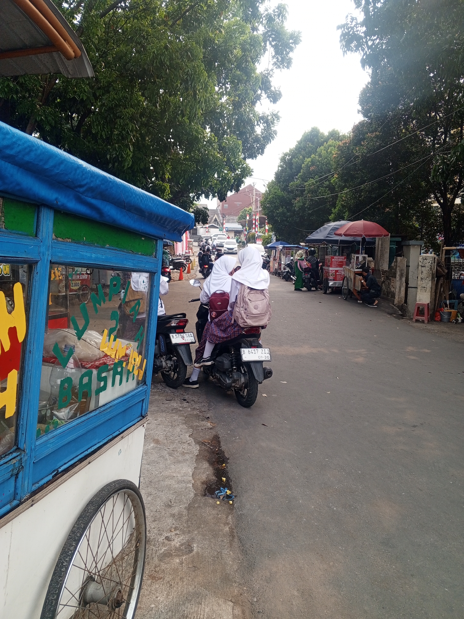 The image depicts a street scene in Depok, Indonesia, with a focus on a mobile food cart in the foreground. The cart is blue and white with a blue tarp covering its top. Yellow and green lettering is visible on the glass of the cart, with "JUMPU" and "BASAH" being partially discernible. A bicycle wheel is attached to the side of the cart. In the background, a paved road runs through a residential area lined with trees. Several people are present, including two young women on a motorcycle, both wearing hijabs and school uniforms, with one riding on the back and carrying a pink backpack. Other individuals are seen near small stalls or shops along the roadside. Motorbikes are parked and in motion on the road. The environment appears to be a typical urban street with a mix of residential and commercial elements.