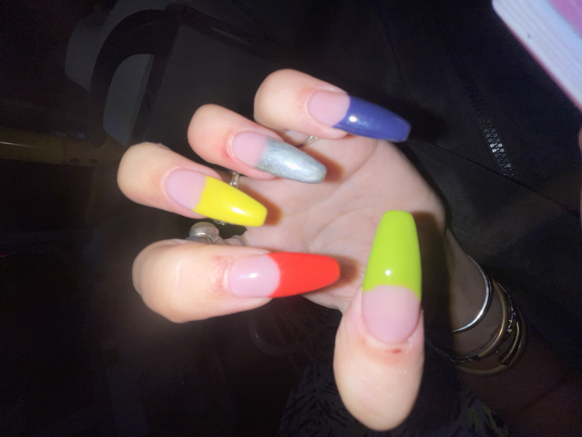 A close-up shot focuses on a person's hand with long, brightly colored, almond-shaped acrylic nails. The nails are painted with a French manicure style, featuring a pink base with a different color tip on each nail. The colors include red, yellow, silver, lime green, and blue. The hand is positioned in front of a dark, out-of-focus background that appears to be an interior setting. There are some rings visible on the fingers. The image does not provide any information or context related to the city of Raanana, Israel.