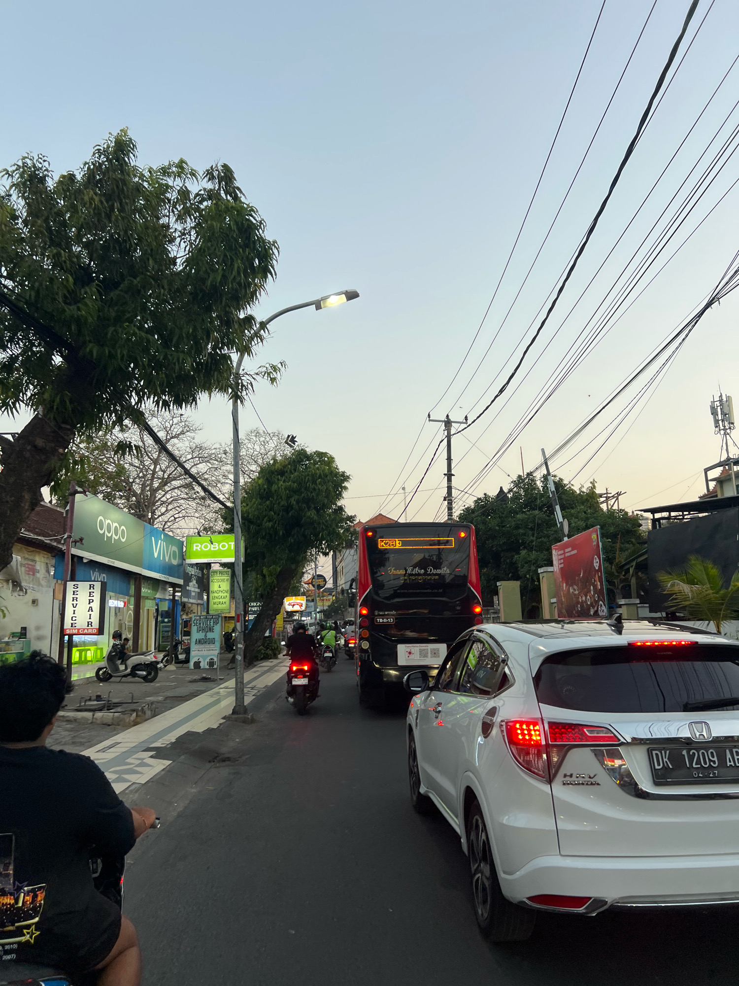 The image depicts a street scene in Kuta, Indonesia, at dusk. A white SUV with license plate DK 1209 AB is in the foreground, with its brake lights illuminated. Behind it, a red and black bus with "Trans Metro Dewata" written on its side is visible. Multiple motorcycles are on the road, some carrying riders. On the left side of the street, shops are lined up, including stores with signs for "Oppo," "Vivo," and "Robot." A lamppost with an illuminated light is present, and overhead power lines crisscross the sky. The sky is a pale orange and blue, indicating the end of the day. A person on a scooter is in the lower left corner of the frame, facing away from the camera.