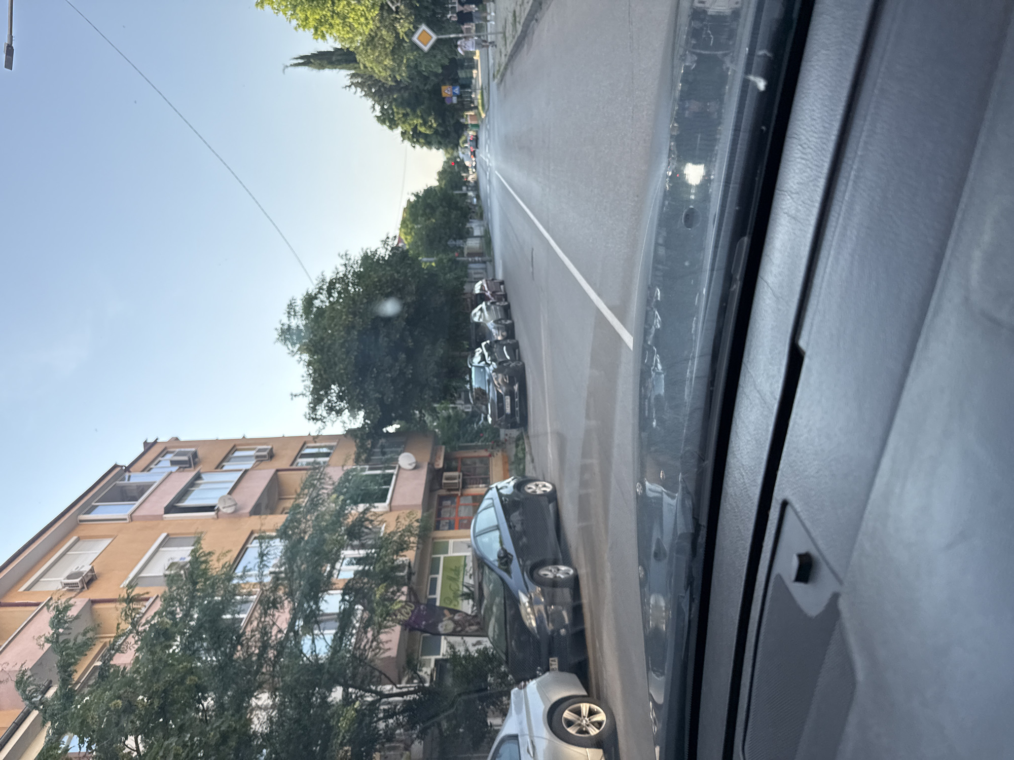 The image captures an urban street scene from the interior perspective of a vehicle, looking forward through the windshield. The upper portion of a dark-colored dashboard, featuring a small rectangular sensor, is visible in the foreground.
The street, identified as being in Stara Zagora, Bulgaria, based on the "STA" license plate prefix visible on a parked vehicle, is an asphalt two-lane road with a dashed white centerline. It extends into the distance, with a red traffic light visible at an intersection.
On the left side of the street, a multi-story building, likely residential, with a peach/orange and pink facade, is partially obscured by mature green trees. Multiple windows, some with external roller blinds, and several air conditioning units are affixed to the building's exterior. The ground floor exhibits commercial storefronts. Several vehicles are parked parallel to the curb on this side, including a silver sedan (partially visible in the immediate foreground), a black third-generation Toyota Prius, and a dark SUV further down.
On the right side of the street, a concrete sidewalk is bordered by green trees, including some taller coniferous species. A yellow diamond-shaped road sign with a white border, indicating a priority road, is visible on the right. Further along the sidewalk, two male pedestrians are observed walking away from the viewer.
The sky overhead is clear and blue, with a faint, thin linear mark, possibly a power line or contrail, crossing diagonally. Sunlight suggests daytime conditions.