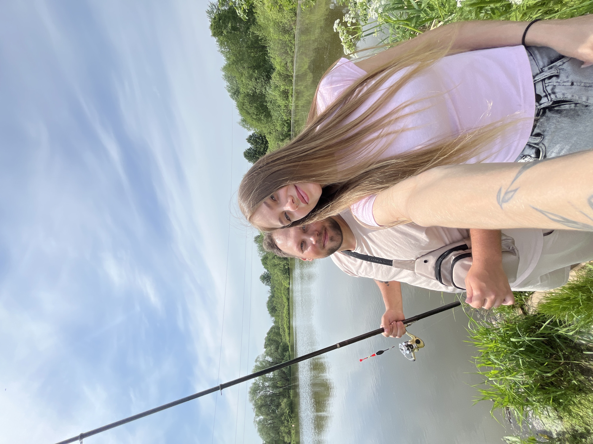 A man and woman are posing for a selfie while fishing in Фоки, Russia. The woman has long brown hair and is wearing a pink shirt and gray pants. The man is wearing a light pink shirt, a tan bag, and light colored pants. He is holding a fishing rod with a bobber. They are standing near a lake with green grass and trees on the bank. The sky is blue with scattered clouds.