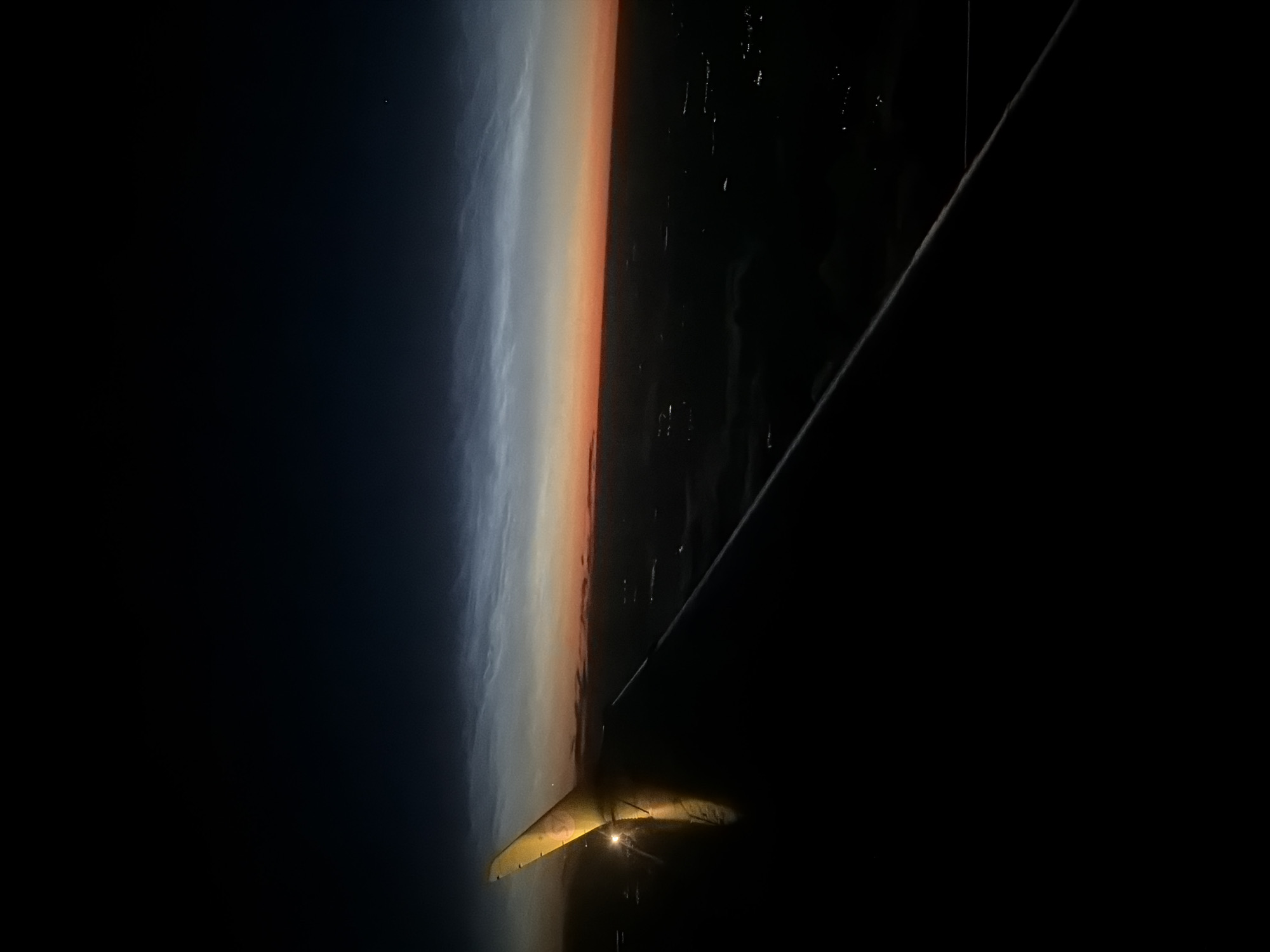 The image is a nighttime aerial view from an airplane window. In the foreground, the dark silhouette of an airplane wing occupies the lower left portion of the frame, with a yellow wingtip catching a bright reflection. The horizon is visible in the mid-ground, displaying a gradient of colors from deep orange at the horizon line, transitioning to pale blue and then to a dark, starry sky above. Wispy, luminous clouds, likely noctilucent clouds, are visible in the upper atmosphere. Below the horizon, a dark landscape is dotted with scattered, bright lights, indicating a populated area. The image does not provide specific details to confirm the location as the city of Ob, Russia.
