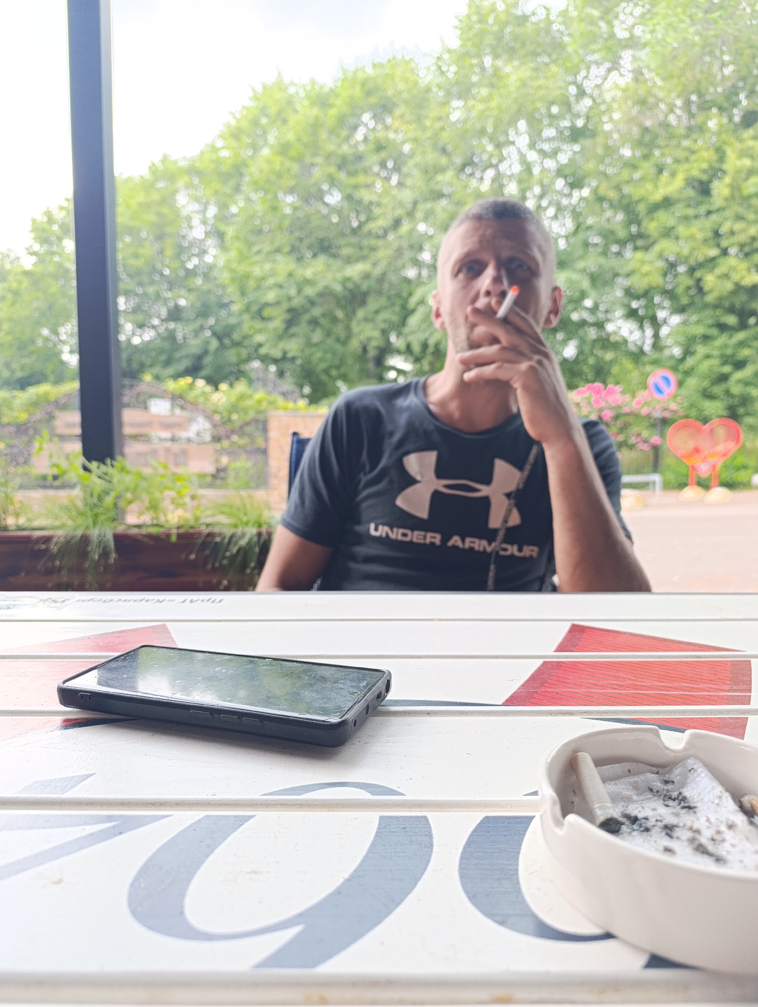 A man is seated at a table, smoking. He is wearing a black t-shirt with the Under Armour logo. In the foreground, a smartphone and an ashtray with cigarette butts are on the table. The scene appears to be outdoors, possibly a cafe or restaurant patio, with trees and foliage visible in the background. The man's gaze is directed away from the camera. There is no indication of the city of Ladyzhyn, Ukraine in the image.