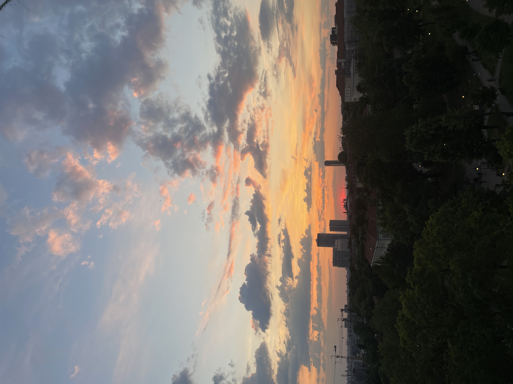 The image captures a sunset view over Vracar Urban Municipality, Serbia. The sky is filled with clouds illuminated by the setting sun, exhibiting hues of orange, yellow, and pink. In the distance, buildings and trees line the horizon. A park with trees and pathways can be seen. Construction cranes are visible in the distance.
