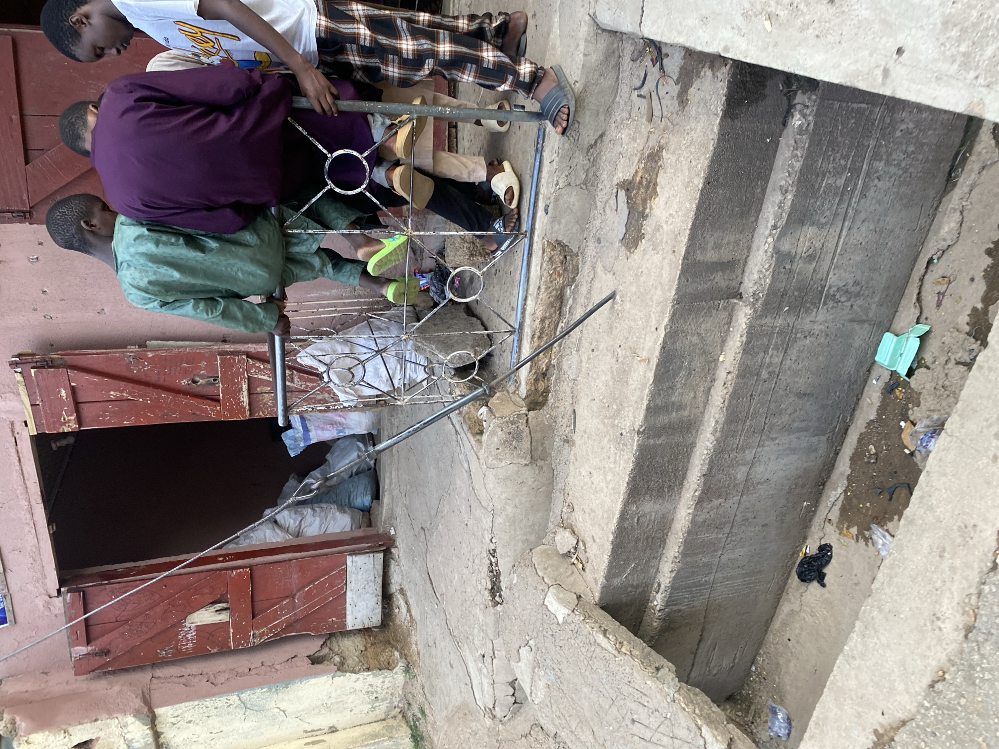 In Jos, Nigeria, the image depicts a scene outside a building with distressed red wooden doors. A group of four young males are standing near the entrance. Three of them are partially visible, with one in a green traditional garment, another in a purple garment, and a third in a white t-shirt with a graphic. They are positioned around a metal railing. Below the railing, the ground is a rough, cracked concrete surface leading to a set of concrete steps. The steps descend into a shallow, open drain or trench. This trench is filled with a small amount of murky water and littered with various pieces of debris, including plastic bags and other refuse. The overall environment appears weathered and somewhat neglected.