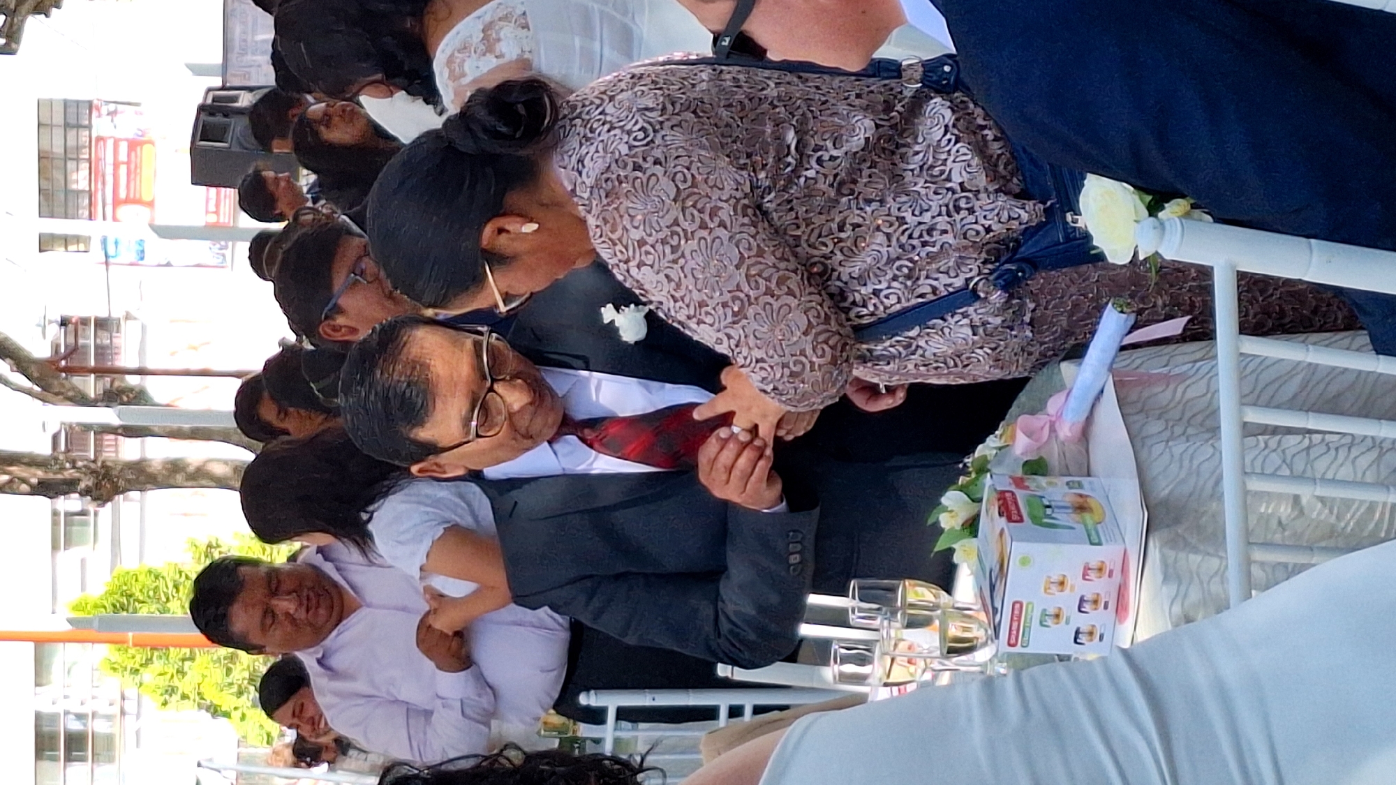 Here is a forensic description of the image:
**Scene:** An outdoor event, likely a wedding, in Cerro Colorado, Peru. Buildings are visible in the background, indicating an urban setting.
**People:** A diverse group of individuals. A man in a suit and tie, wearing glasses, is having a ring placed on his finger by a woman with her hair in a bun. Other people are standing around them, observing.
**Objects:** A white chair, decorated with a white rose and tied flowers. There is a box with food items on the chair, as well as multiple glasses of what appears to be champagne.
**Actions:** The focus is on the exchange of rings between the man and woman. Other individuals are likely participating in or observing the event.
**Environment:** The setting is outdoors, with buildings in the background. There appear to be trees or plants nearby. The event seems to be taking place during the day.