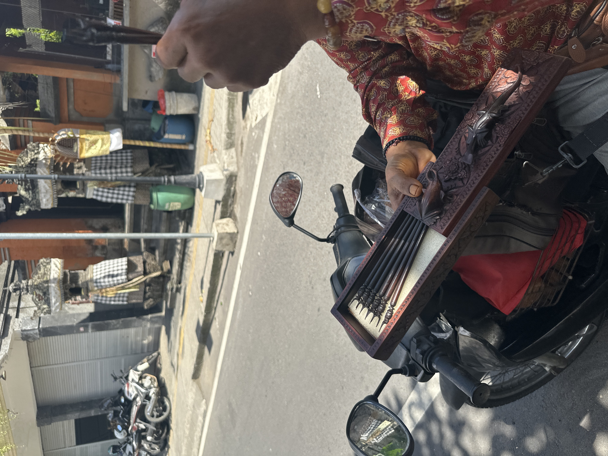 In Ubud District, Indonesia, a person is sitting on a motorcycle, holding a wooden box filled with carved wooden items. The box is intricately carved with animal motifs. The person is wearing a red patterned shirt and jewelry on their wrist. In the background, there are buildings with traditional Indonesian architecture, several motorcycles parked on the side of the road, and green and blue plastic barrels. The scene appears to be a street in a bustling urban area.