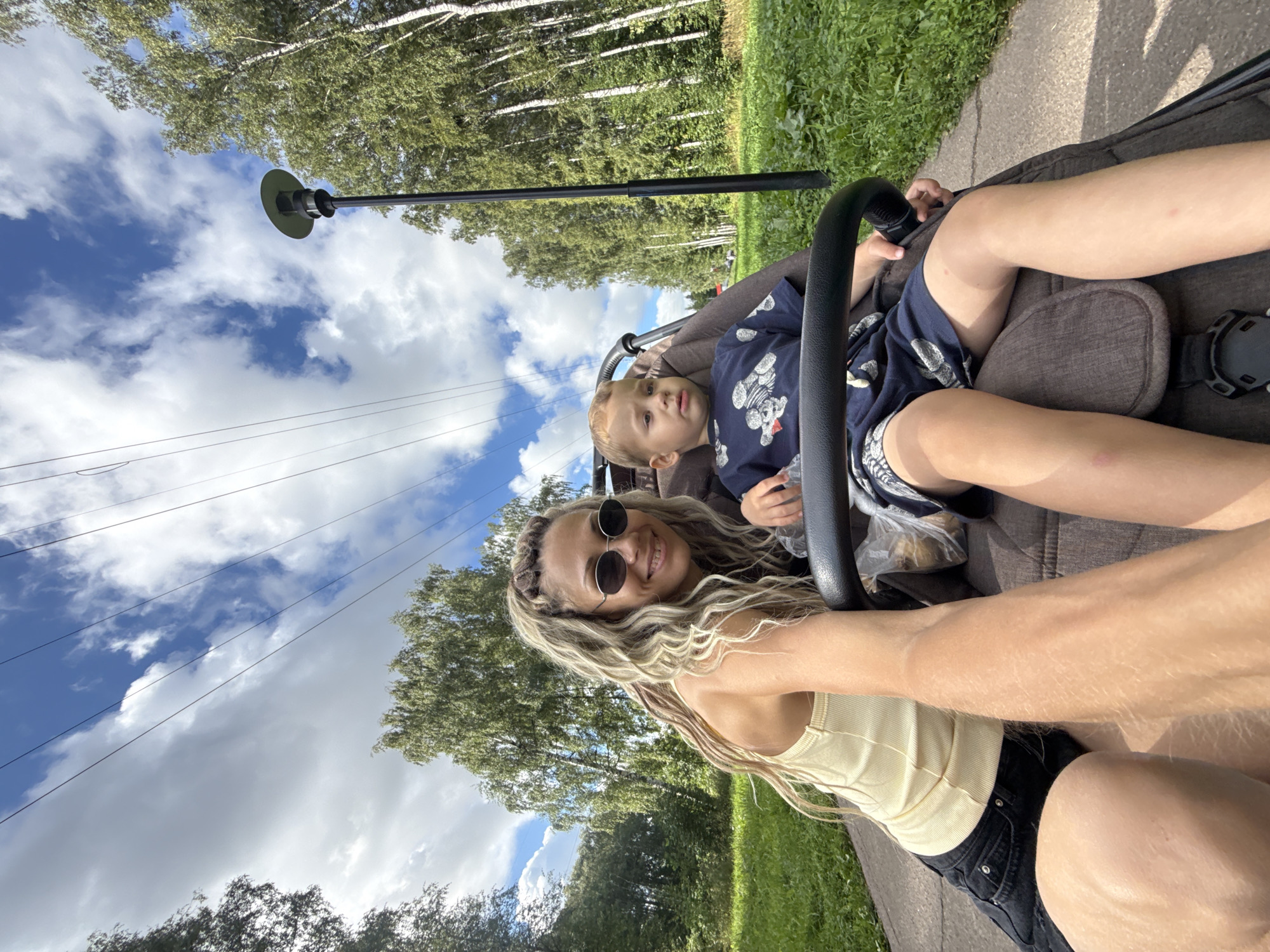 In the city of Rusanovo, Russia, a woman and a young child are seated in a stroller on a paved path. The woman, with long blonde dreadlocks and wearing sunglasses and a yellow tank top, is on the left, leaning towards the camera with her arm extended. She is smiling. The child, in a blue t-shirt with a pattern of bears, is seated to her right. The stroller is brown. To the right of the path, a tall, modern street lamp stands amidst a dense forest of birch trees. Overhead, the sky is a vibrant blue with scattered white clouds and crisscrossing power lines. The overall scene is outdoors, during the daytime, in a park-like setting with abundant greenery.