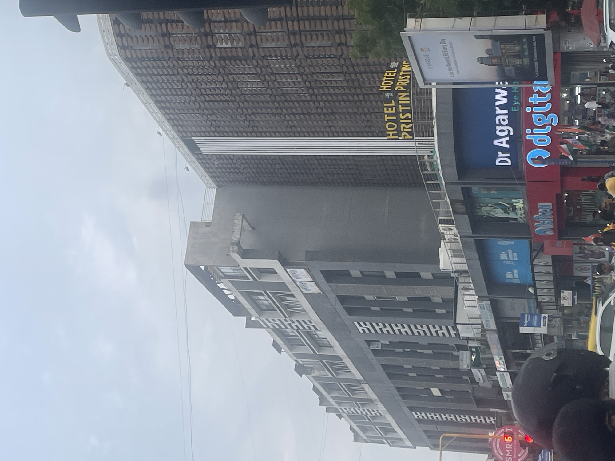 This is a medium-shot, eye-level, vertical, outdoor photograph taken on a cloudy day in Sarkhej, India. The image depicts a multi-story building complex with a modern architectural style. The building on the right features a distinctive facade with a textured, patterned design and vertical white stripes. A large sign with the text "HOTEL PRISTIN PRISTINE" is prominently displayed on this building. Below this hotel, there are shops and businesses. One sign reads "Dr Agarwal Eye Hospital," and another displays the word "Odigita" in large, illuminated letters. There are also shops with signage such as "Alifa." Several Indian flags are visible, suggesting a national holiday or celebration. In the foreground, parts of two people wearing helmets are visible, indicating they are likely on a motorcycle or scooter. Other people are seen on the street level, and cars are present in the lower portion of the image. Traffic lights are also visible. The overall scene is a street view of a commercial area in Sarkhej, India.