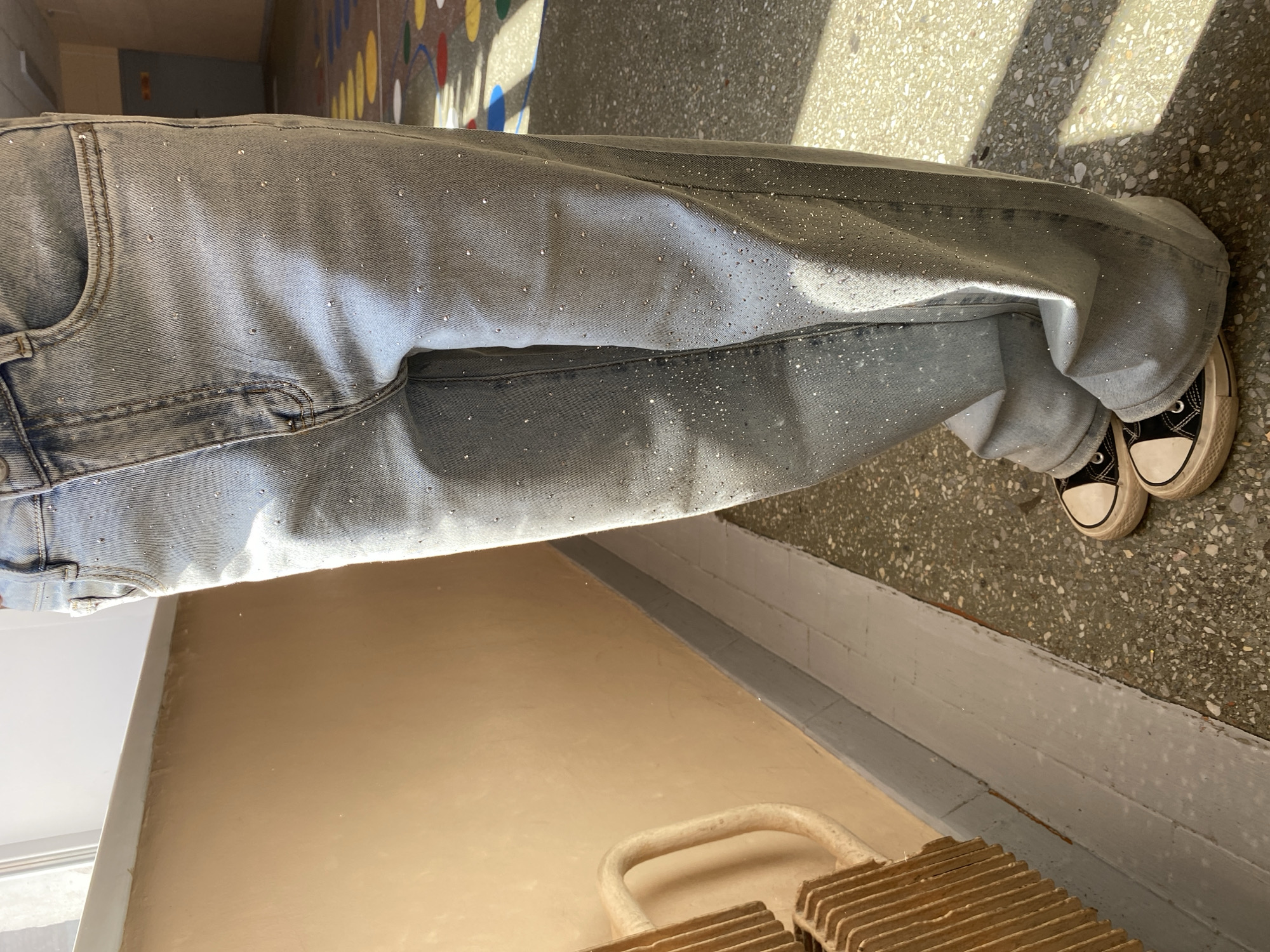 The image shows a person wearing light-wash, wide-leg jeans adorned with small, reflective embellishments. They are also wearing black and white canvas sneakers. The person stands indoors on a concrete floor, and part of a beige-painted wall is visible behind them. A radiator sits near the wall. It is not possible to determine the location in the city of Votkinsk, Russia.
