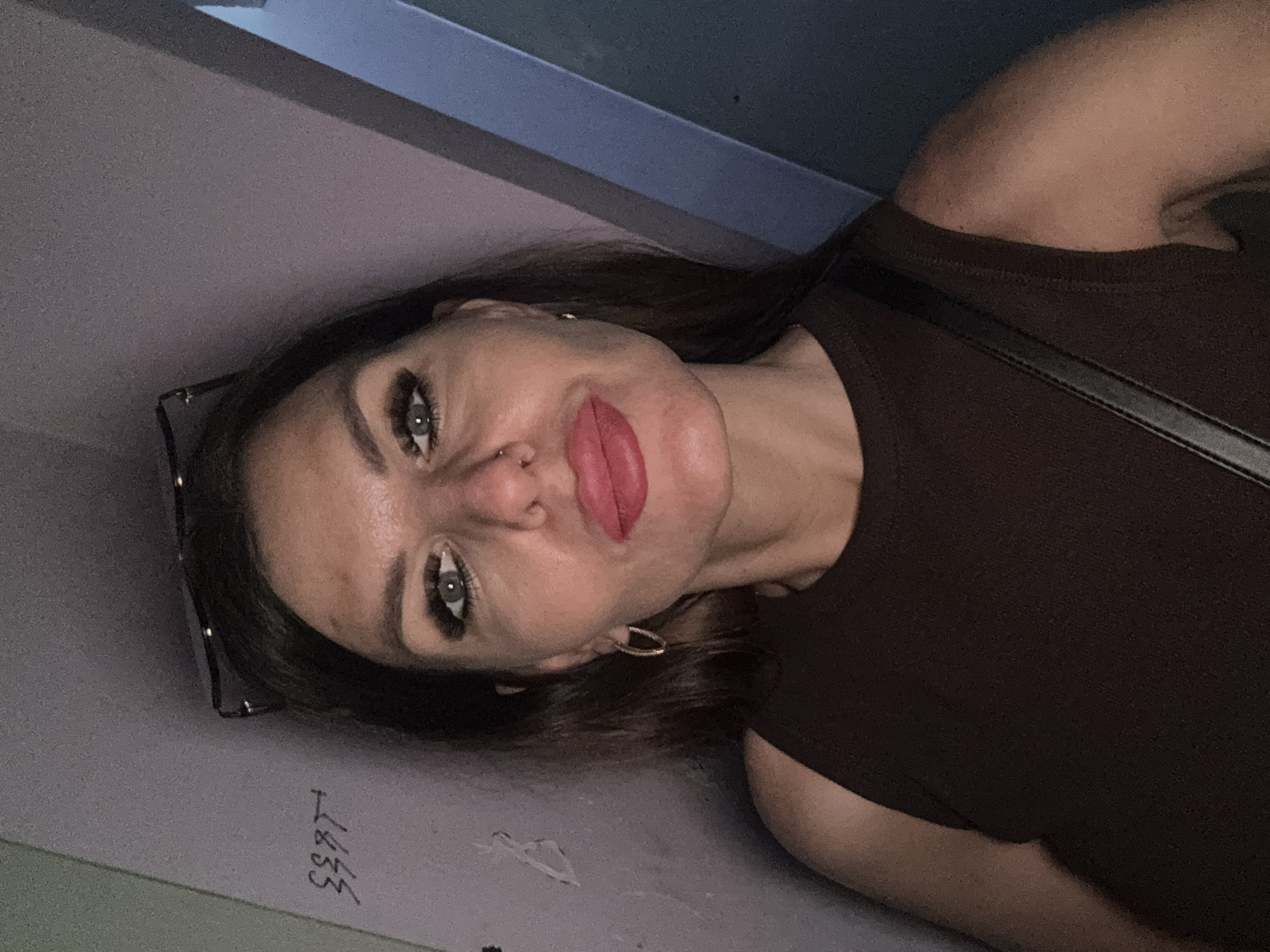 A woman is photographed indoors. She is wearing a brown sleeveless top and has dark hair, light eyes, and red lipstick. She has a nose piercing and is wearing hoop earrings. Glasses are placed on the left side of the image next to her face. The wall behind her is gray with some markings written on it. The background suggests an interior location. No specific landmarks or details indicate the location is Chaikovsky, Russia.