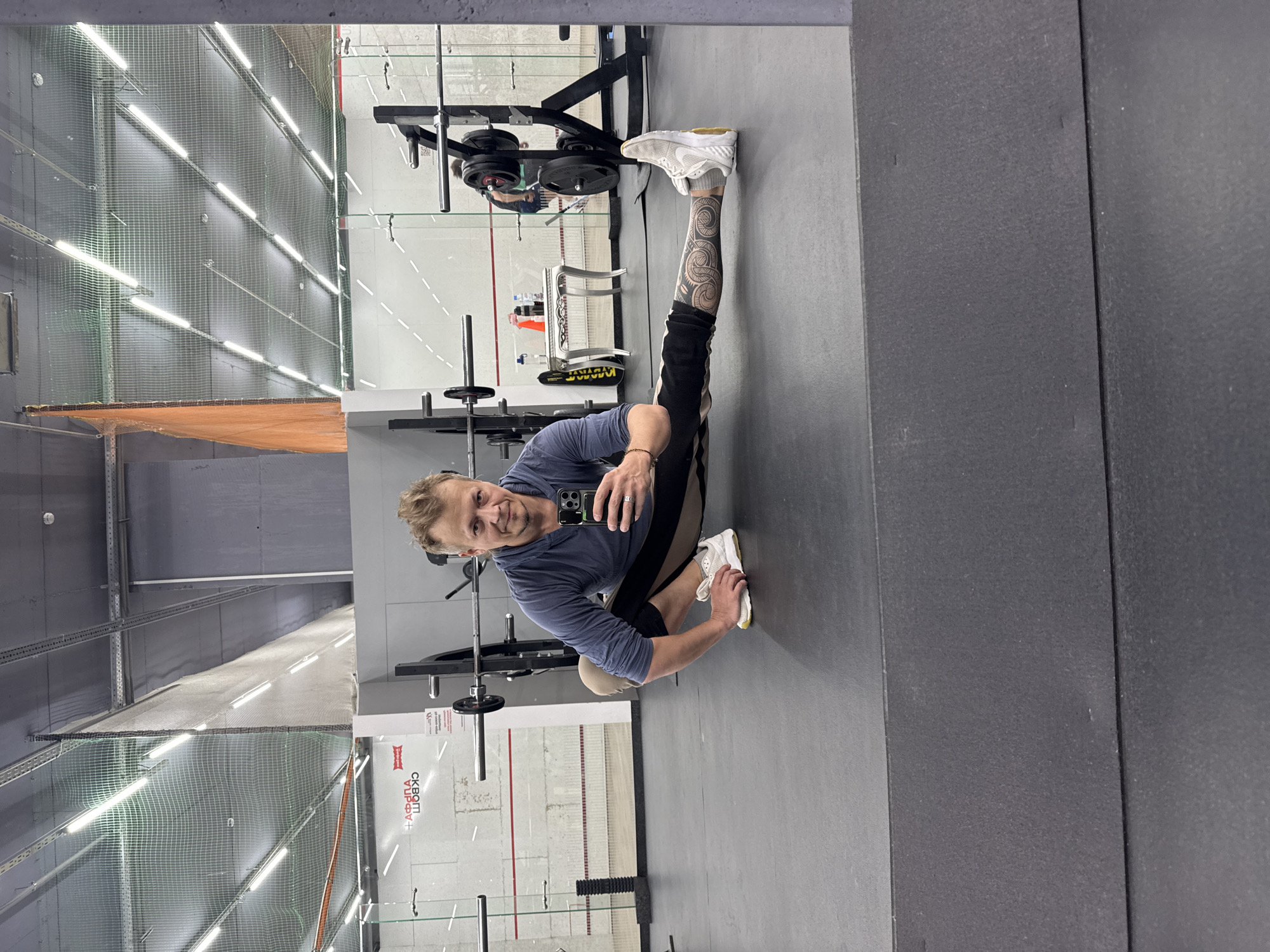 A man is visible in a selfie taken in the mirror of what appears to be a gymnasium in the city of Novosibirsk, Russia. He is squatting, holding a smartphone in his hand, looking at the camera with a smile. He is wearing a long-sleeved shirt and black pants, and his right leg has tattoos on it. The gym equipment visible includes weightlifting racks and bars. The gym also has squash courts lined up against the walls. The building’s interior is illuminated by overhead fluorescent lighting. A partially obscured orange safety net can be seen in the background.