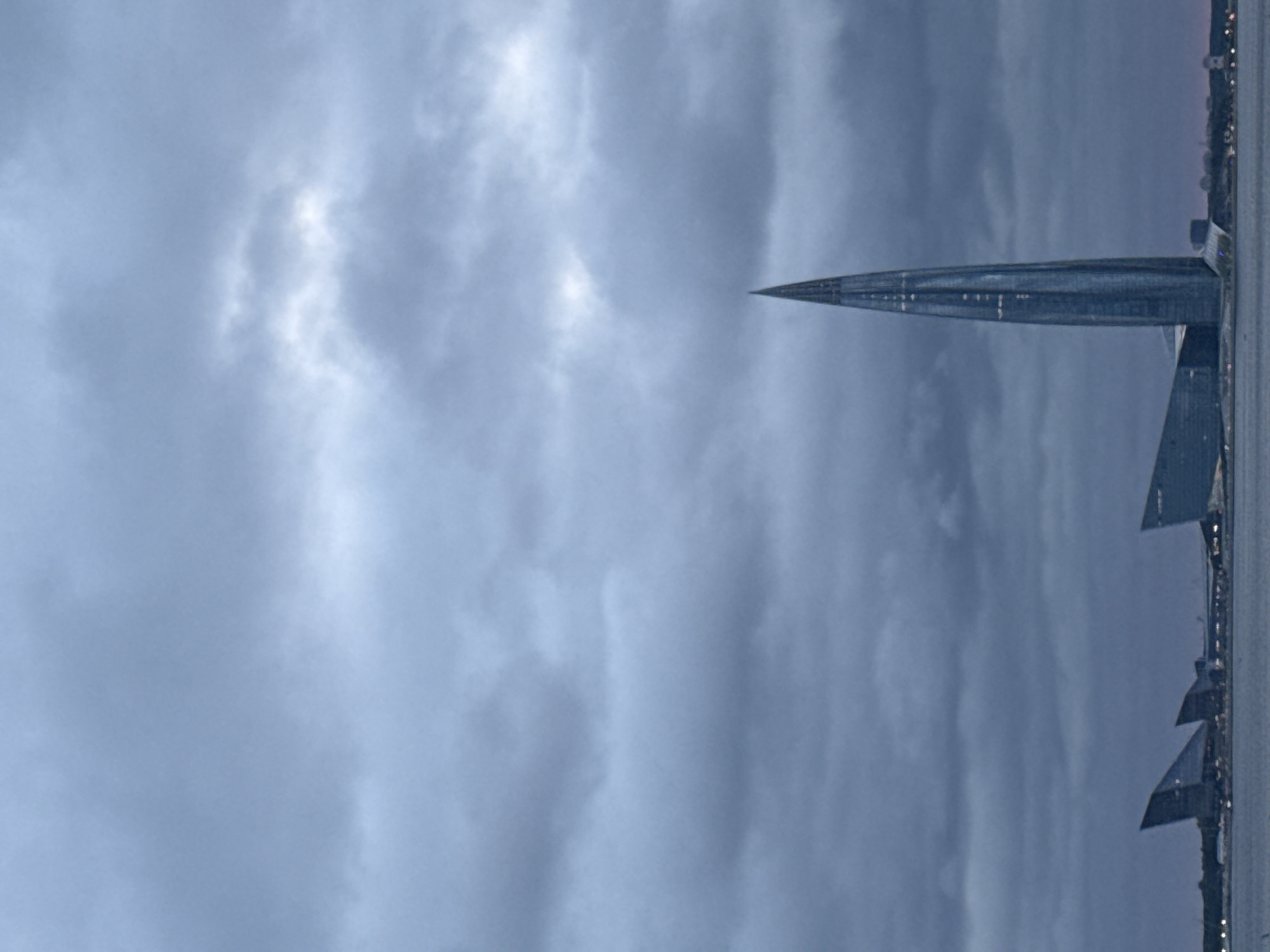 The image captures the Lakhta Center, a modern skyscraper located in the Primorsky District of Saint Petersburg, Russia. The tall, needle-like structure dominates the skyline, its glass facade reflecting the overcast sky. To the left of the skyscraper, the curved, angular forms of other buildings, possibly part of the same complex or nearby architectural structures, are visible. The scene is set against a backdrop of heavy, dark clouds, suggesting inclement weather or twilight. A body of water, likely the Gulf of Finland, stretches across the bottom of the frame, with the lights of a distant cityscape twinkling along the shoreline. No people or specific actions are depicted. The overall impression is one of a modern urban landscape under a dramatic sky.