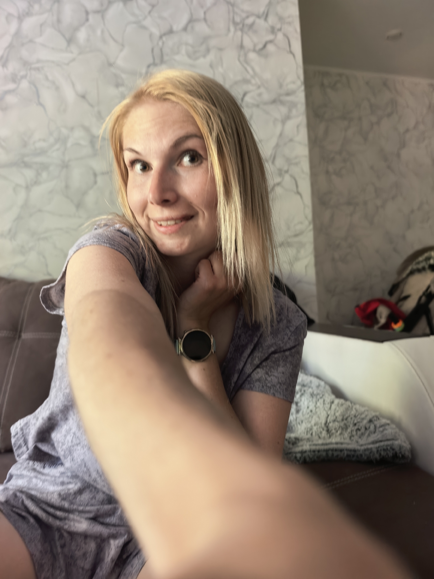 A blond woman is taking a selfie while sitting on a brown couch. She is wearing a gray t-shirt and shorts and has a smartwatch on her left wrist. A white wall with a marble pattern is behind her. Another white couch with items on it is to the right of the brown couch. The location is likely indoors. The city of Sterlitamak, Russia cannot be confirmed from the image.