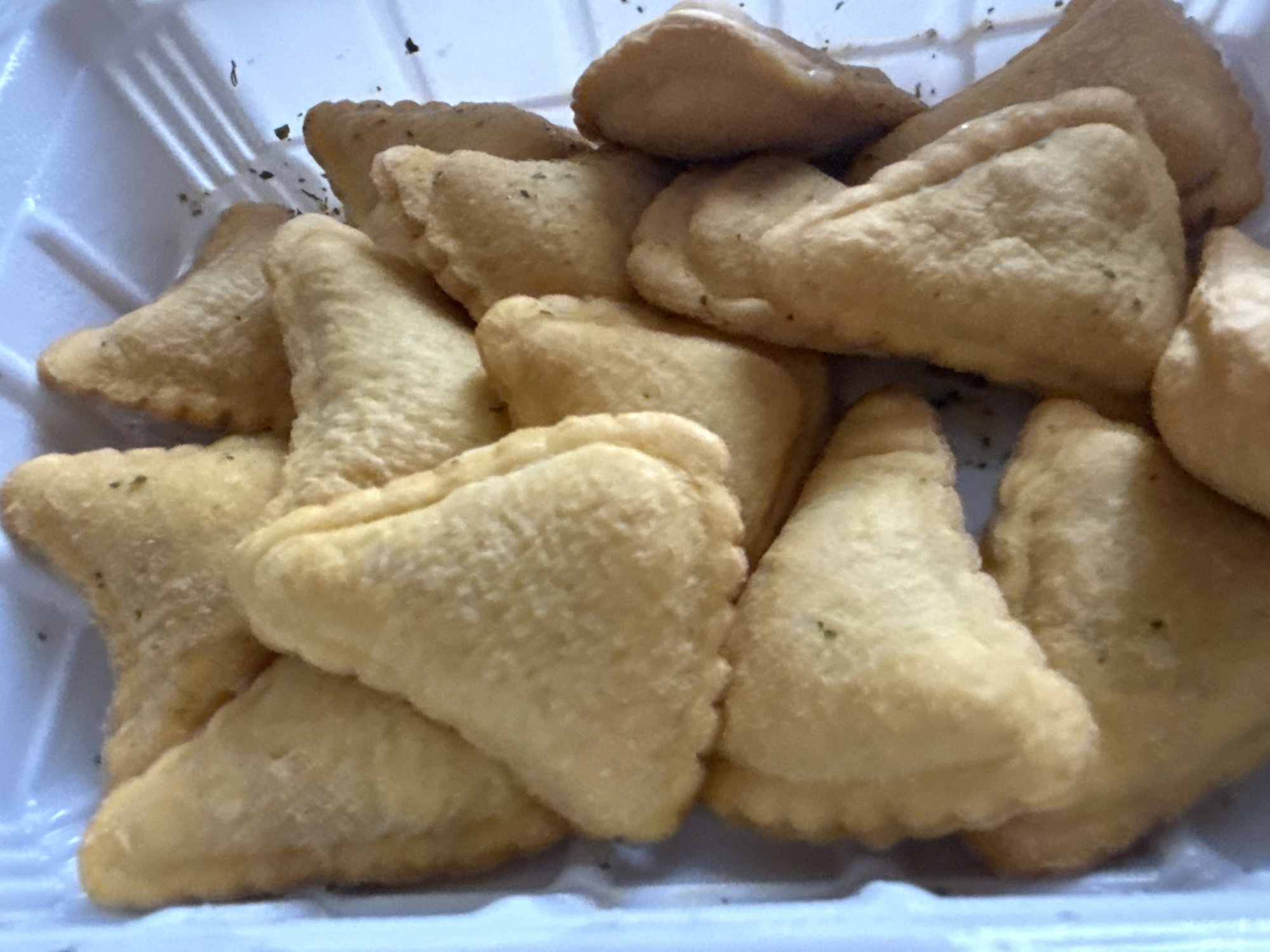 The image shows a close-up of a pile of fried ravioli-like pasta in a white plastic container. The pasta pieces are irregularly shaped, roughly triangular, and golden brown in color. A small amount of green seasoning is visible on the surface of some pieces. The container appears to be made of polystyrene.