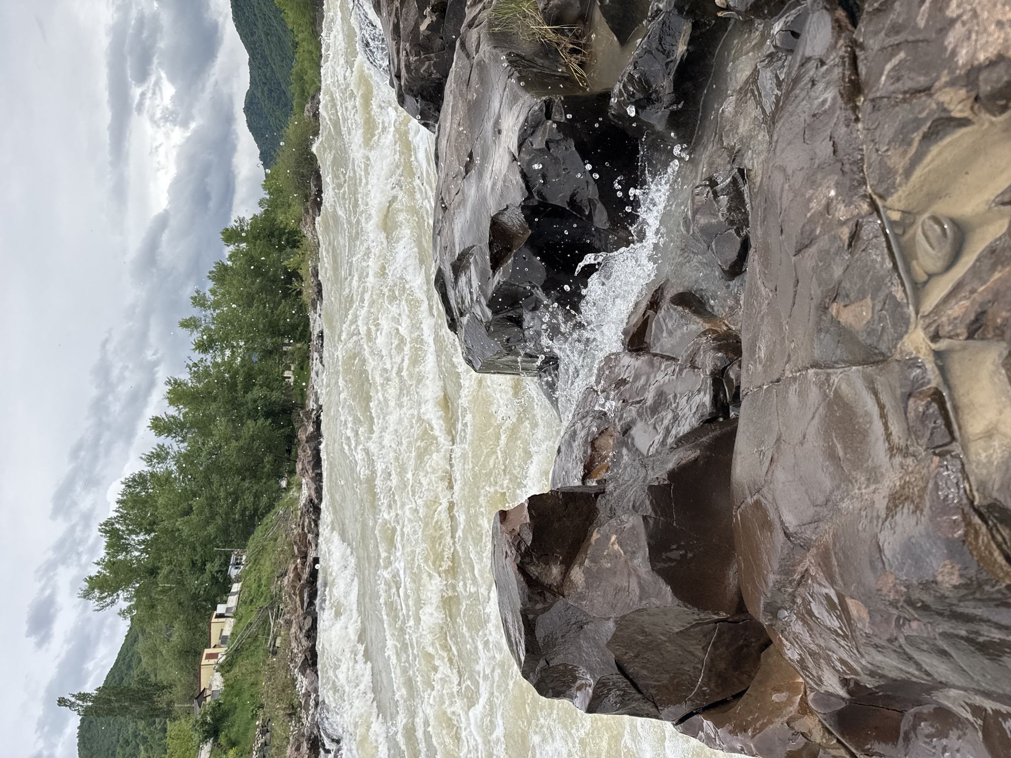 The image depicts a fast-flowing river with turbulent, white-capped water. The river is flanked by large, dark, wet rocks in the foreground, with water splashing on them. On the left bank, there is a line of trees and some buildings. The sky is overcast with grey clouds. The location is Каменномостский, Russia.