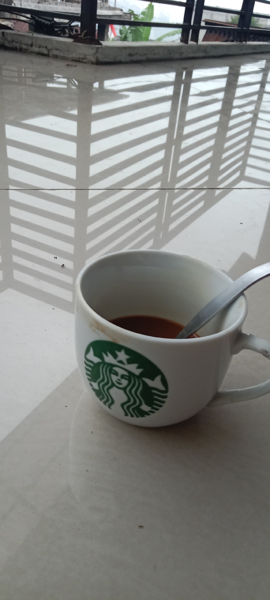 A white Starbucks mug, containing a dark liquid, rests on a light-colored tiled surface. A metal spoon is partially submerged in the liquid. The mug has a brown stain near its rim and the iconic green Starbucks logo is visible on its side. The tiled surface reflects the shadow of a striped structure, possibly a railing or a blind, which casts a pattern of diagonal lines. In the background, a concrete ledge supports some vegetation, including a large green leaf. The scene appears to be an outdoor setting. The image was taken in the city of Krinjing, Indonesia.