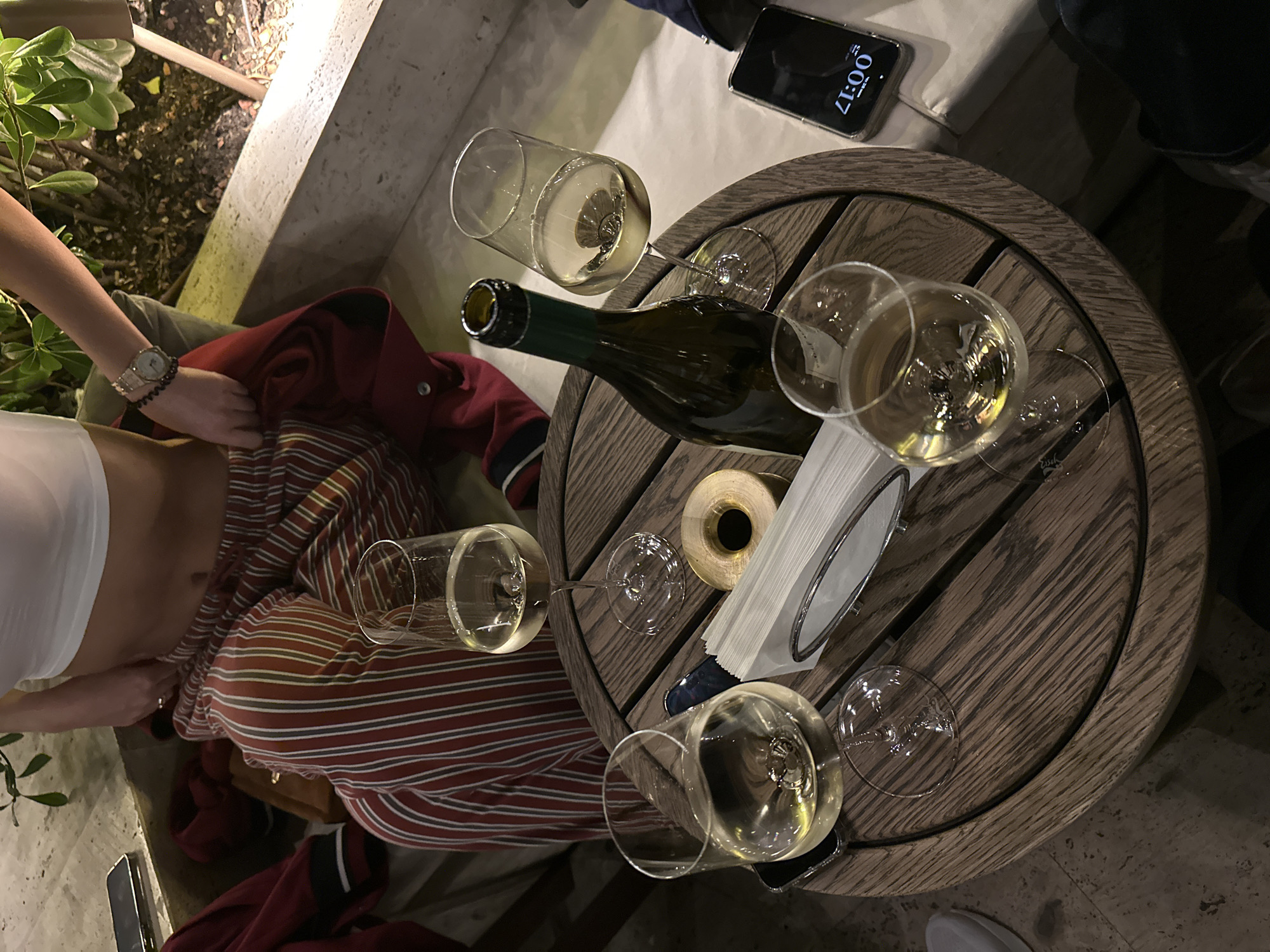 The image shows a top-down view of a wooden table, presumably outdoors. On the table are a wine bottle, four wine glasses filled with white wine, a roll of tape, and a napkin holder with napkins. A smartphone with the screen displaying a time of 00:17 is on the table, near the upper right corner. Part of a person is visible on the left side, wearing a white top and striped pants, with a red jacket nearby. Based on the style and ambiance, the location might be an outdoor dining area or patio in Tbilisi, Georgia.