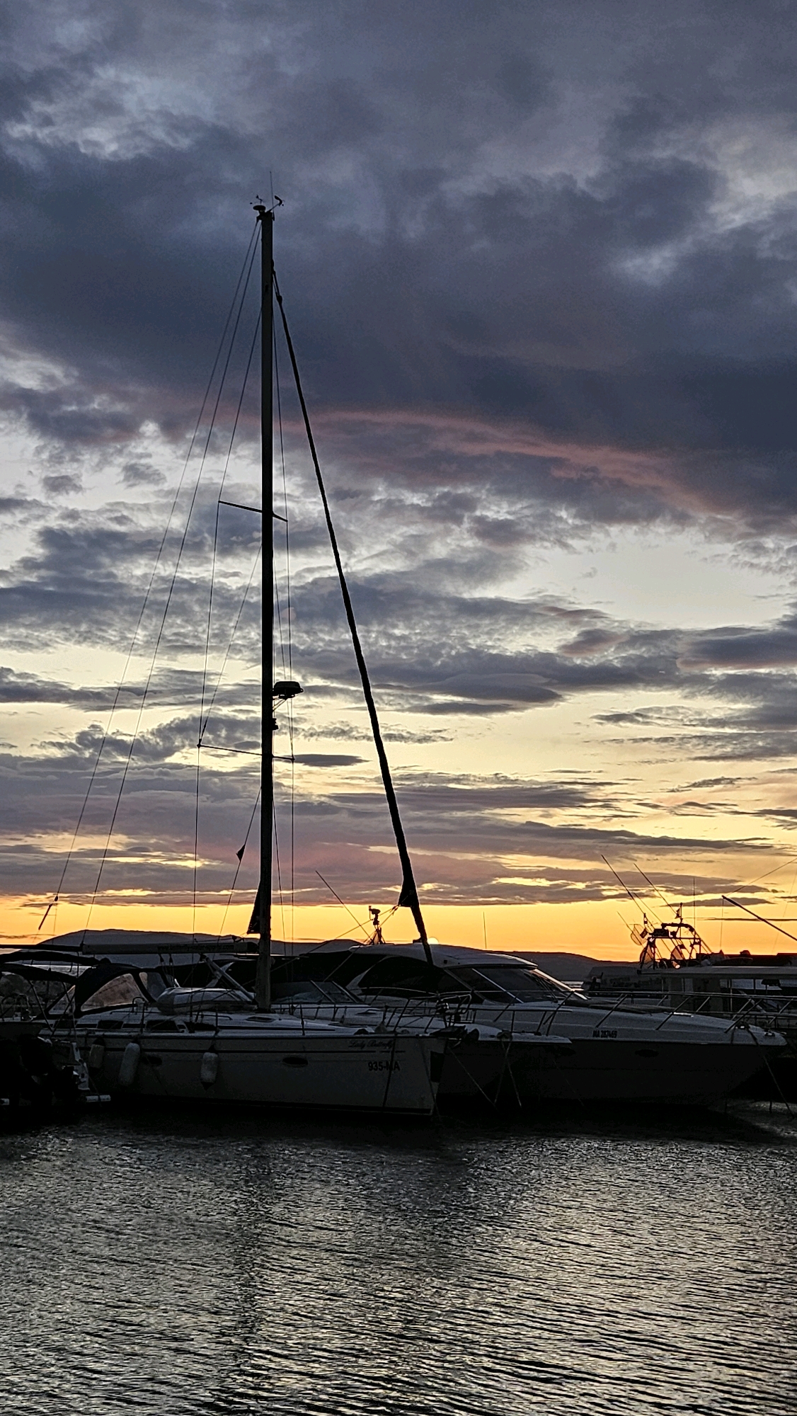 The image depicts a marina at dusk in Općina Tučepi, Croatia. A sailboat with its mast prominently silhouetted against a dramatic sky dominates the center of the frame. The sky is filled with a gradient of colors, transitioning from dark, brooding clouds at the top to hues of orange and yellow near the horizon. Several other boats, including what appear to be motor yachts, are docked alongside the sailboat. The water is dark and reflects the ambient light. In the distance, a silhouetted mountain range can be seen. The scene suggests a tranquil evening in a coastal town.