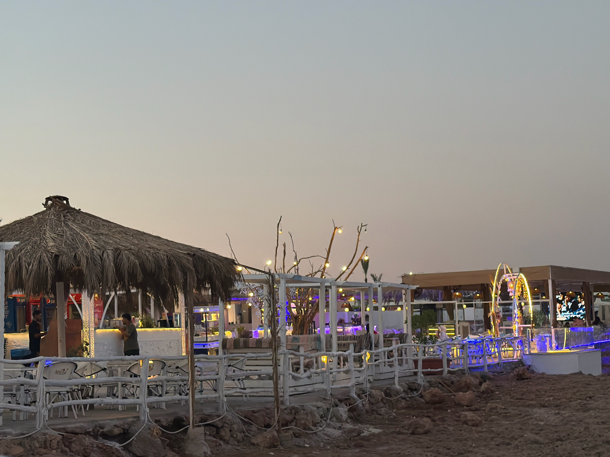 This image depicts a beachfront dining area in Sharm El Sheikh, Egypt, likely during dusk or early evening. The scene features a thatched-roof structure on the left, resembling a bar or open-air eatery, with tables and chairs set up outside. Further to the right, a more modern open-sided structure is illuminated with string lights and blue neon lights, creating a vibrant atmosphere. Decorative branches with lights are also visible. The ground in the foreground is sandy and appears somewhat unkempt. In the background, there are glimpses of other structures and lights, suggesting a developed tourist area. Two individuals are visible, one standing near the thatched-roof structure and another in the distance near the illuminated seating area. The sky is a pale gradient of blue and orange, typical of twilight.