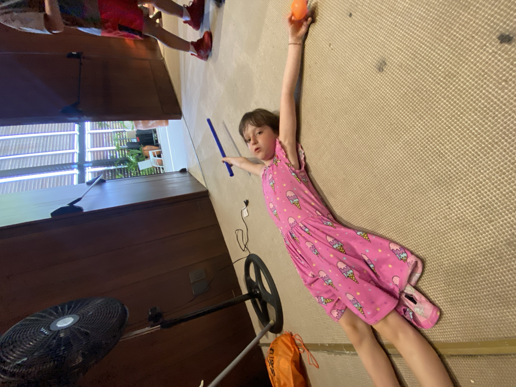 The scene appears to be indoors, likely within a residence. A young girl wearing a pink dress with an ice cream cone pattern is lying on a tan carpeted floor. She holds a blue cylindrical object in one hand and an orange ball in the other. To the left, there is a dark wooden structure, possibly a cabinet or wall. A standing fan is partially visible in the lower left corner. Another child's legs are visible in the upper left corner. An orange bag is near the fan. The location in the city of Marina Maroma, Mexico is not discernible from this image.