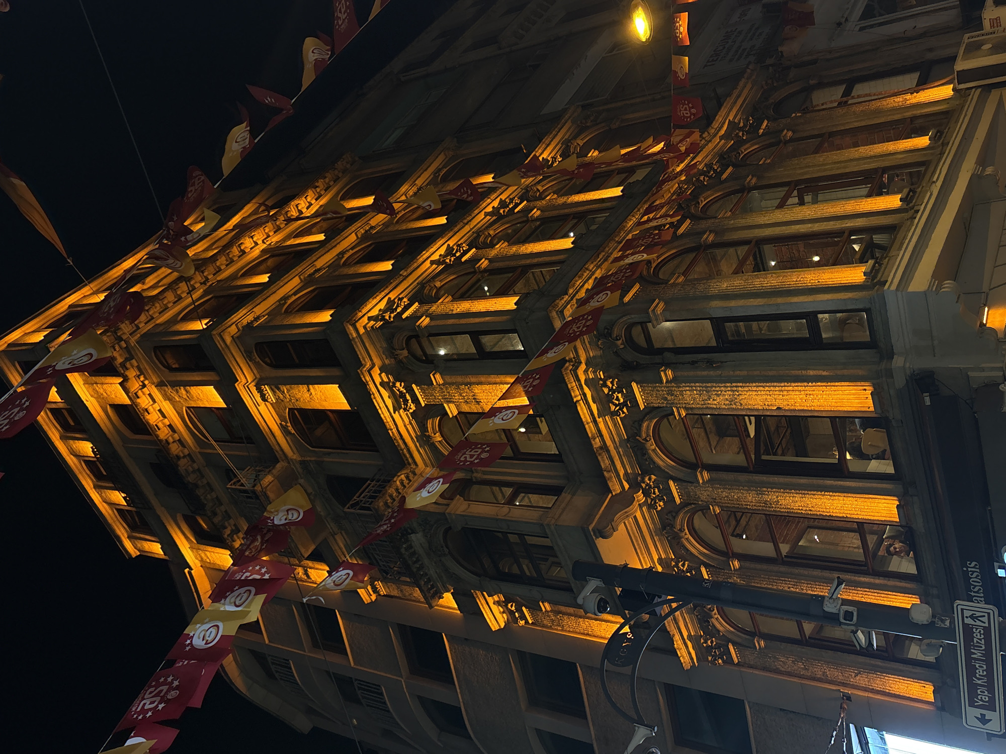This is a nighttime photograph capturing a richly detailed building in the Beyoğlu district of Turkey. The multi-story structure dominates the scene, illuminated by warm, golden-orange uplighting that accentuates its ornate architectural features, including numerous arched windows, decorative columns, and cornices. Red and yellow flags, many displaying the letters "GS" and numbers such as "25" or "26," are strung in garlands across the street and between buildings, prominent in the foreground and extending into the background.
On the lower left facade of the main building, a white street sign with a left-pointing arrow reads "Yapı Kredi Müzesi" (Yapı Kredi Museum) and partially "atsosis." A dark utility pole with a security camera is positioned in the lower-left foreground. Through one of the large arched windows on the lower level, a person is visible looking outwards. To the immediate left of the main building, a less ornate, lighter-colored building with rectangular windows is partially visible. On the far right, a street light emits a bright yellow glow. Further to the right, a sign partially reads "TERCÜME" (Translation), and air conditioning units are mounted on a lower facade. The sky is dark, indicating night.