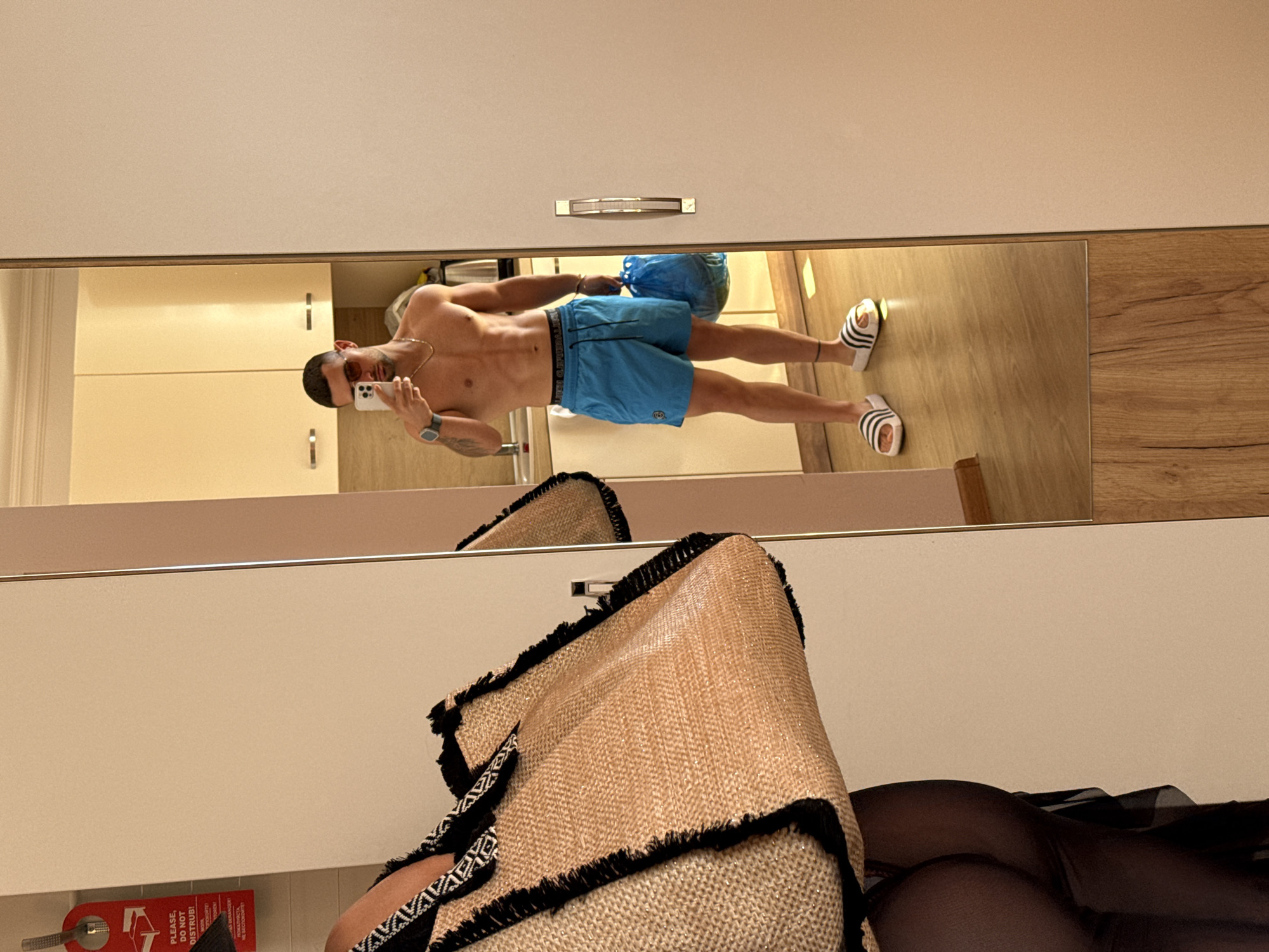 A man with a muscular physique stands in front of a mirror, taking a selfie. He is wearing blue swim trunks, sunglasses, and white Adidas slides. He is holding a blue plastic bag in his right hand and a phone in his left. In the reflection, his body is visible from the waist up, and he appears to be in a hotel room. Behind him, a kitchen counter with cabinets is visible. The room's flooring is light wood. The "Do Not Disturb" sign is hanging on the door. The image was taken in Nesebar, Bulgaria.