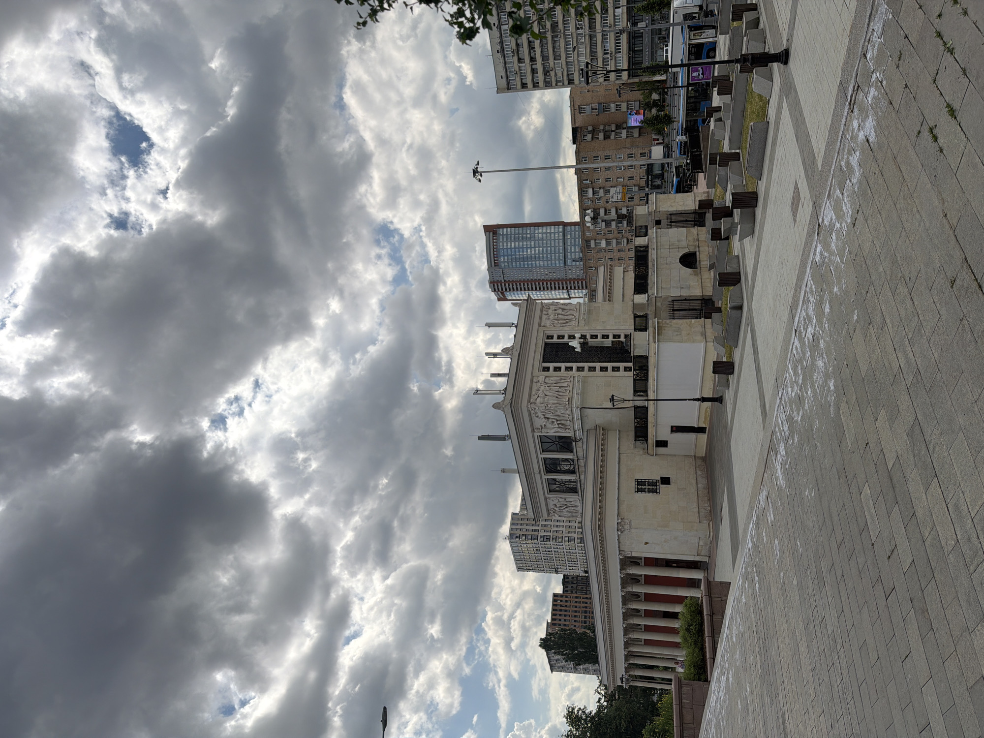 This is an eye-level shot of a neoclassical building with a colonnade and ornate frieze under a cloudy sky. In the background, several modern high-rise buildings are visible. A paved plaza with planters and benches is in the foreground. A bus is visible on the street behind the plaza. The overall scene suggests an urban environment with a mix of old and new architecture.
