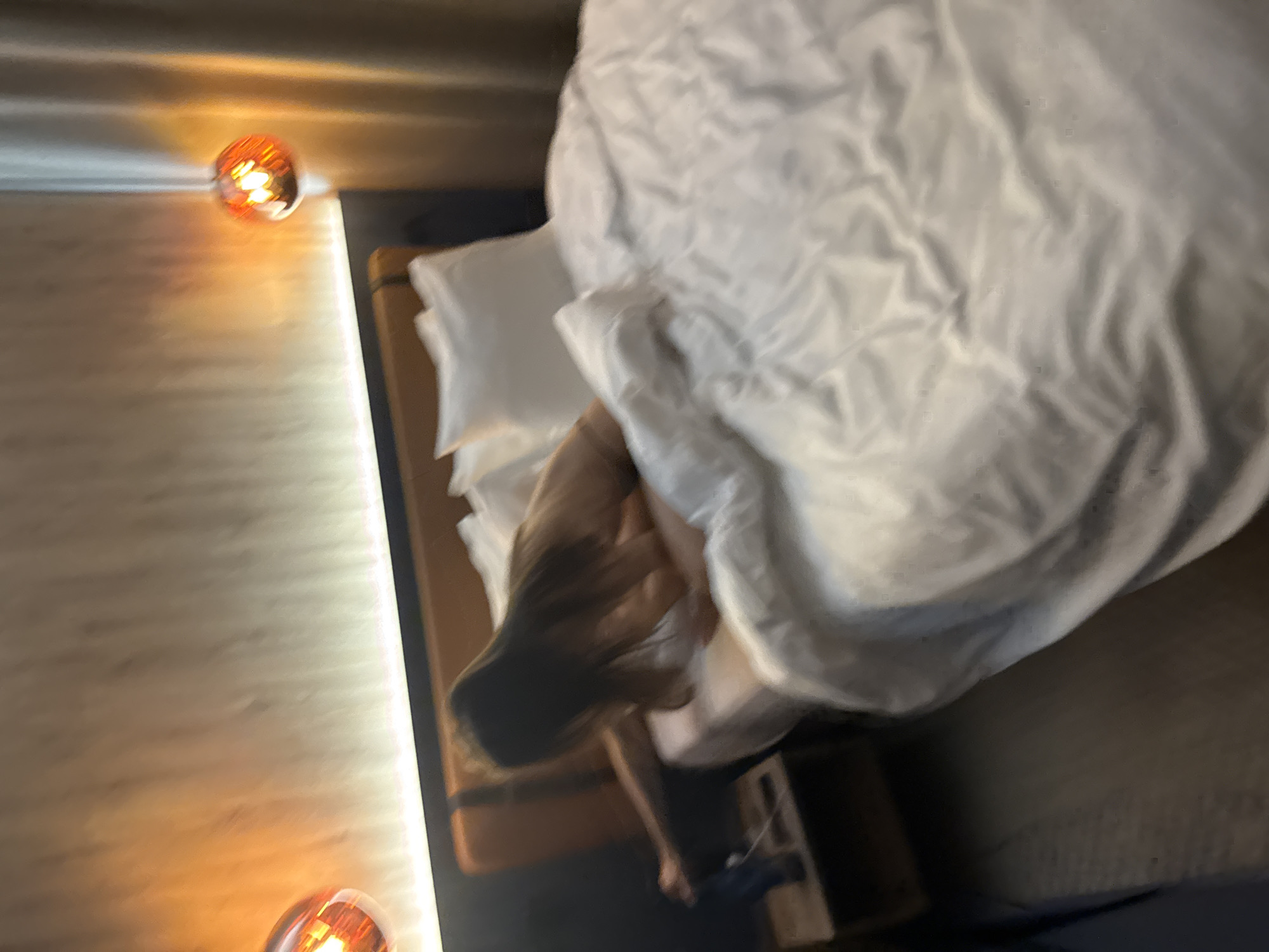The image presents a blurry, overhead, and tilted view of a bedroom scene. A person, appearing to be female with long, dark hair, is partially visible in the bottom left portion of the bed, covered by a rumpled white duvet or blanket. Her hair is spread out, suggesting movement, such as stretching or shifting while in bed.
The bed itself features white pillows and a large white duvet. To the left of the bed, a wall with subtle horizontal texturing is illuminated by a vertical strip light. Two spherical light fixtures, emitting a warm orange or amber glow, are mounted on this wall, one in the top left and another in the bottom left corner of the frame. A wooden surface, possibly part of the bed frame or a bedside table, is partially visible near the person. The foreground and bottom right of the image show a dark surface, likely the floor or carpet.
The activity appears to be a person resting or just beginning to stir in bed. The warm artificial lighting from the fixtures suggests an indoor setting, possibly during the evening or night, contributing to a calm and intimate atmosphere. There are no clear location cues beyond a typical bedroom setup, possibly a hotel room or a modern home. No text is visible in the image.