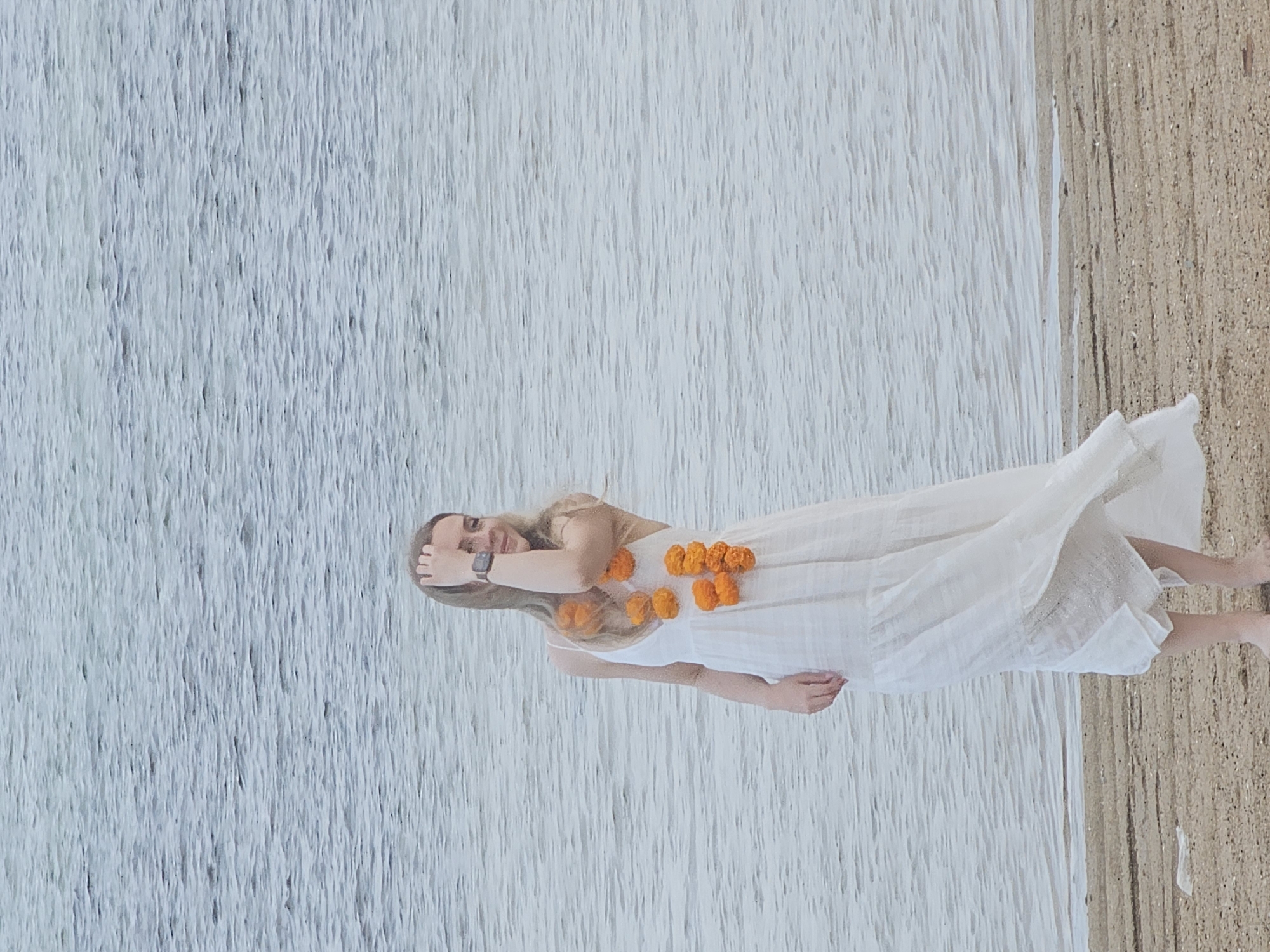 A woman in a white dress adorned with an orange flower garland around her waist stands on a beach. The water, with visible ripples, laps at the shore beside her. The sand appears light brown and textured. The woman is looking towards the camera with a relaxed expression, her hand resting on her head. A smartwatch is visible on her left wrist. The scene is brightly lit, suggesting daytime. The water is a light blue-gray, and the sky is not visible. The overall impression is one of peacefulness and natural beauty.