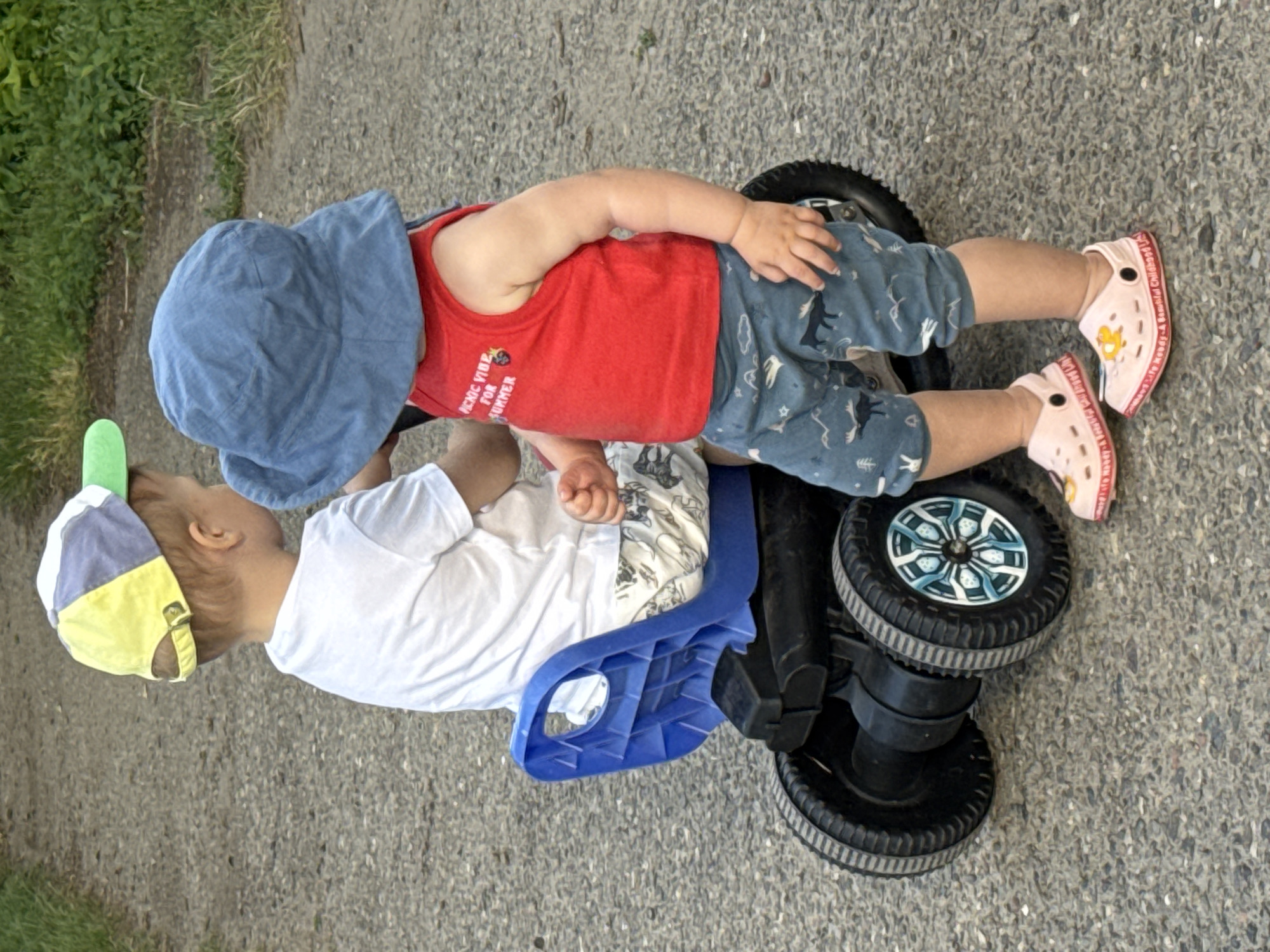 In Minusinsk, Russia, two young children are captured in a photograph. One child, wearing a white t-shirt and a multicolored baseball cap, is seated in a blue plastic toy car. The other child, in a red tank top and a blue sun hat, stands beside the car. Both children are wearing patterned shorts. The ground appears to be a paved pathway with some grass and foliage visible in the background. The image is taken from a medium distance, focusing on the children and the toy car.