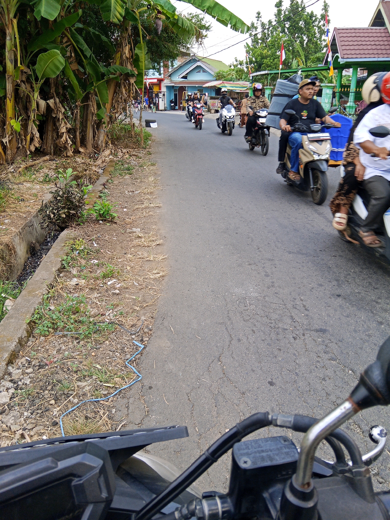The image depicts a street scene in Lubuklinggau, Indonesia, with several individuals riding motorcycles. On the right side of the frame, a person wearing a white shirt and black pants rides a motorcycle, their lower legs and feet visible. To their left, another individual is seated on a motorcycle, wearing a black t-shirt and blue jeans, carrying a large grey item on the back. Further down the road, more motorcycles are seen, some with passengers. The environment includes a paved road with a concrete drainage ditch on the left, lined with dry grass and some vegetation. Banana trees with large leaves are prominent on the far left of the image. Buildings with blue and light-colored walls and red-tiled roofs are visible in the background, along with flags and electrical wires. The perspective suggests the photo is taken from the viewpoint of someone on a motorcycle.