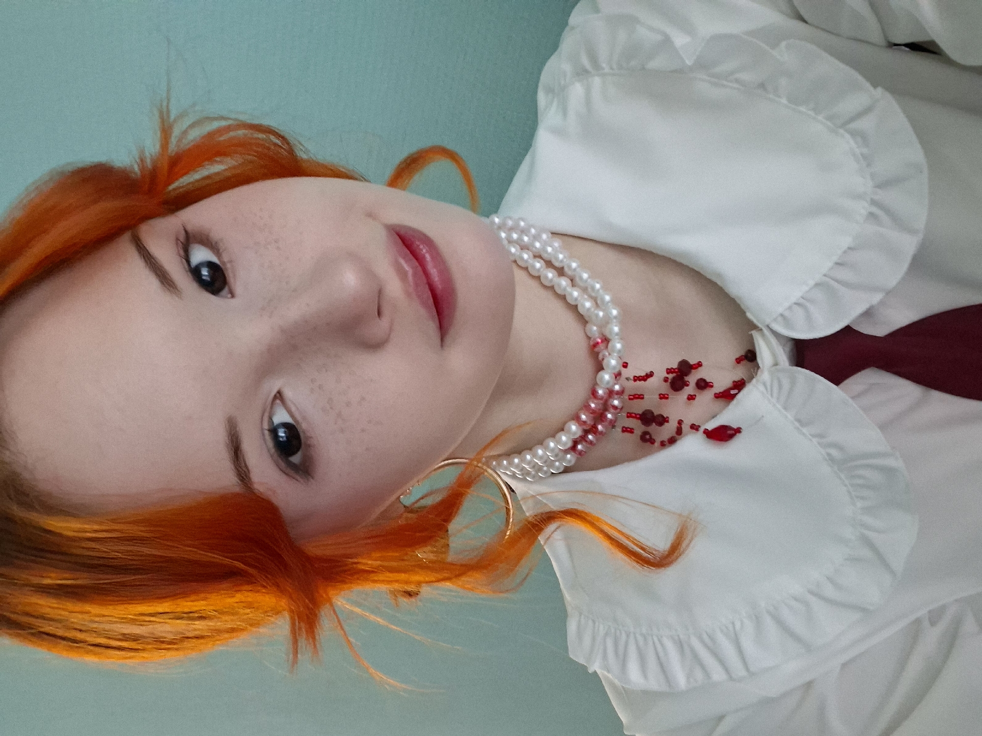 A rotated portrait-style image captures a young woman with vibrant, bright orange-red hair. She has fair skin, prominent freckles scattered across her nose and cheeks, and dark eyes, gazing directly at the viewer with a soft, engaging smile. Her lips are adorned with red lipstick.
She is dressed in a white blouse featuring ruffled details around the collar and sleeves. Layered around her neck are two necklaces: one a double strand of white pearls, and the other a delicate arrangement of small red beads. A dark red tie or scarf is visible beneath the collar of her blouse. A gold hoop earring can be seen on her left ear. The background is a plain, solid light blue-teal color, indicating an indoor setting. The lighting appears soft and even, illuminating her features. No visible text is present. The scene is set in Chaikovsky, Russia.