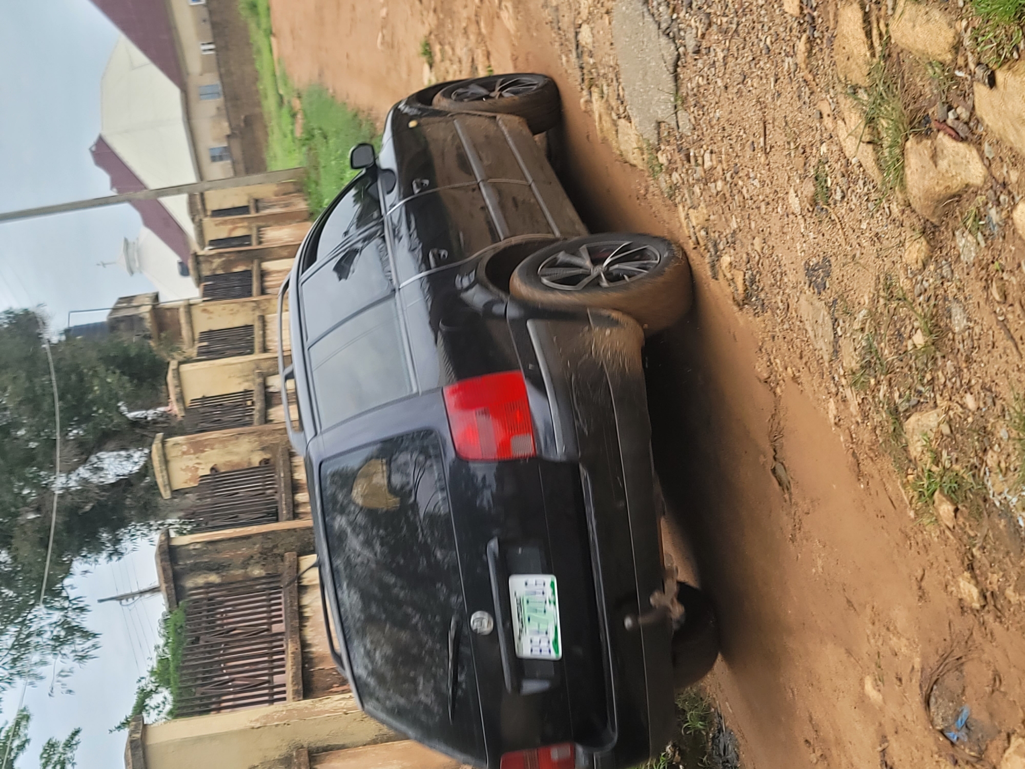 In Barakin Akawo, Nigeria, a black Volkswagen Passat station wagon is parked on a dirt road. The car, which appears to be dusty, has a license plate that reads "B58-774D NG." A weathered fence with vertical metal bars runs along the side of the road, with a cream-colored wall and pillars. In the background, a church with a white dome and red-tiled roofs is visible. The sky is overcast.
