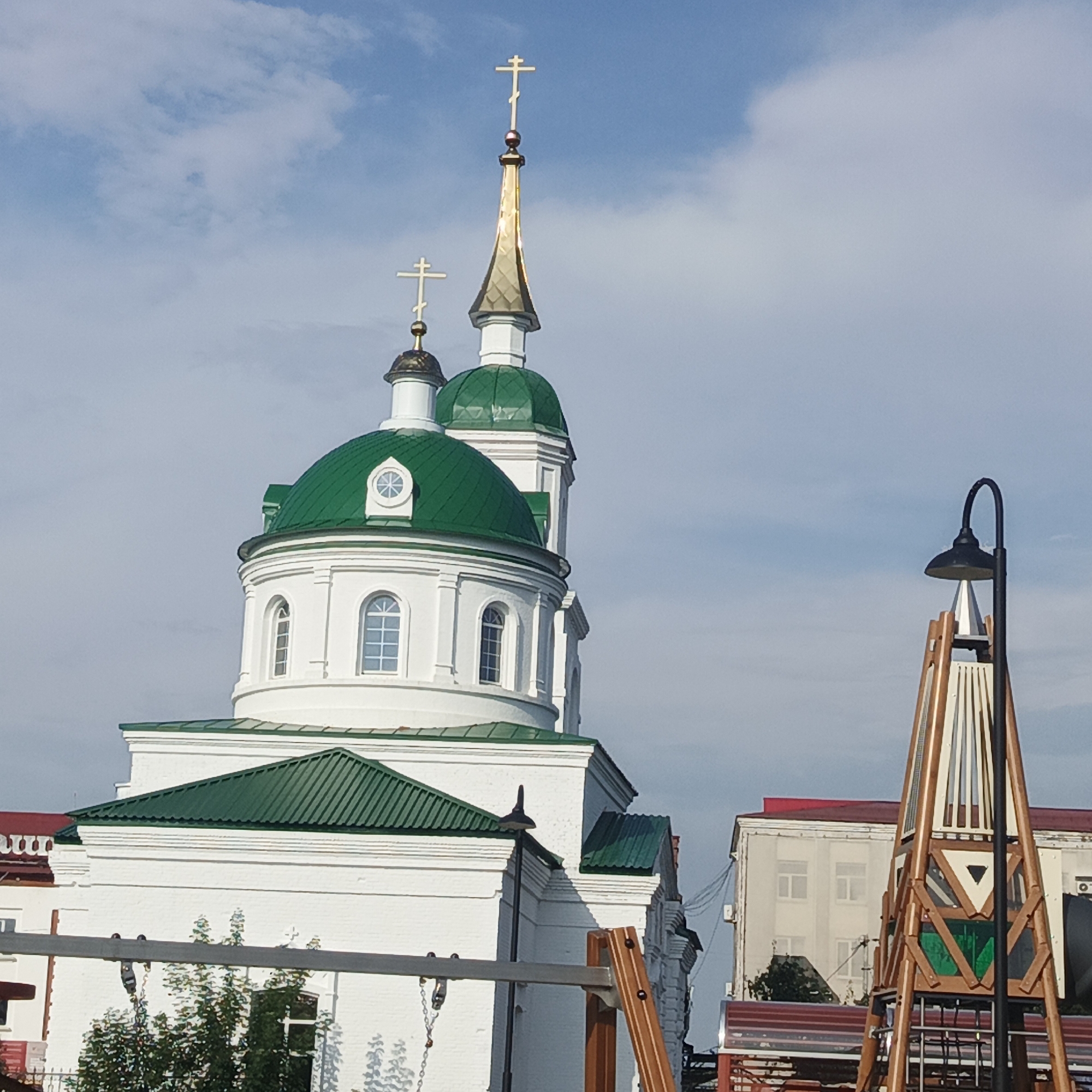 The image shows a white church with green domes and crosses against a blue sky with white clouds. In the foreground, there is a modern, wooden playground structure with a black lamppost attached to it. To the right of the church, there is a light-colored building with a red roof. The church is located in Kambarka, Russia, and is identifiable by its unique architectural features. There are no people visible in the image.