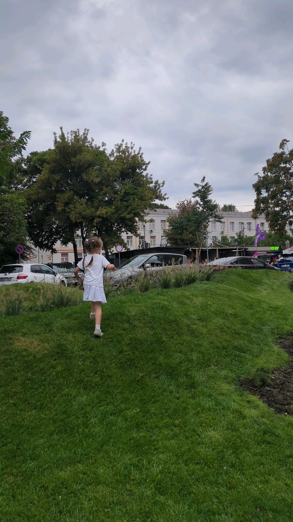 A young girl with blonde braided hair, wearing a white t-shirt and white skirt, runs across a grassy hill. In the background, cars are parked along a street lined with trees. Beyond the street, a multi-story building with white walls and dark-trimmed windows is visible. The sky is overcast with gray clouds. The scene appears to be in a park or public area within the city of Chișinău, Moldova, as evidenced by the architecture and general urban landscaping.
