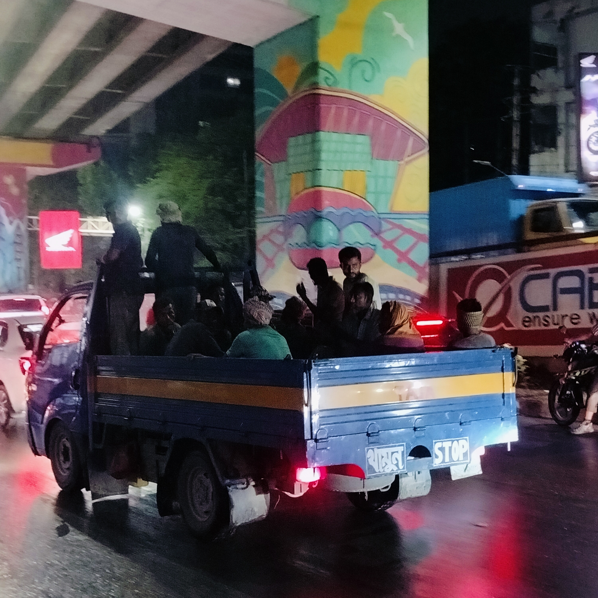 The image captures a blue truck transporting a group of people in Dhaka, Bangladesh, at night. The truck bed is filled with several individuals, some standing and others sitting. The truck's rear displays a "STOP" sign in English, along with text in Bengali. The scene appears to be under an overpass, evidenced by the overhead structure and vibrant mural on the support columns. Signage, including a red Honda sign, is visible in the background, along with other vehicles and a pedestrian on a motorbike, all contributing to the urban environment. The street appears wet, reflecting the city lights and contributing to the overall ambience.
