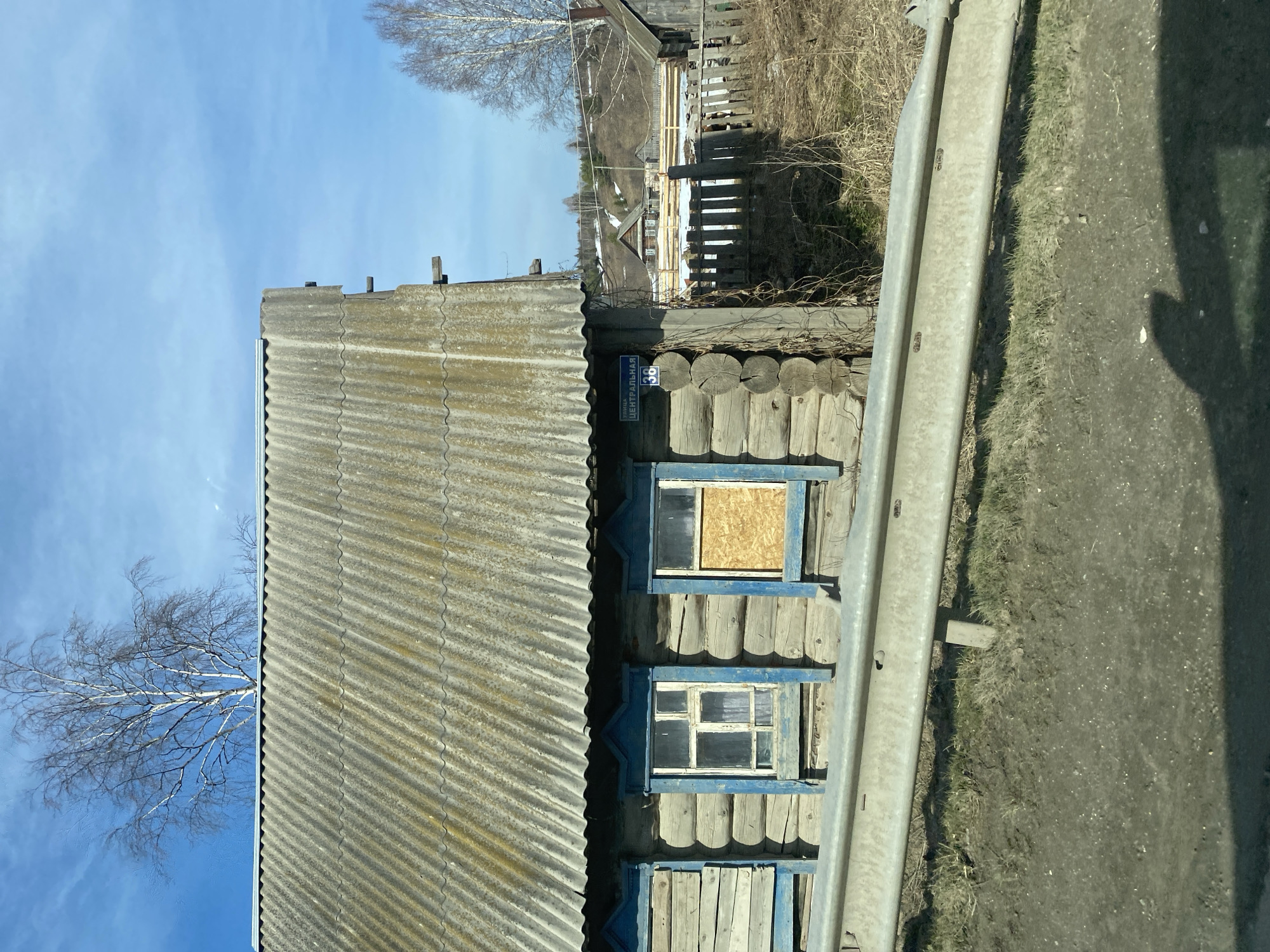 A weathered, log cabin sits adjacent to a road in Verkh-Usa, Russia. The house, possibly number 36 based on a sign, features a corrugated roof and blue-trimmed windows. One window is boarded up. A metal guardrail runs along the road beside the house. The environment includes a bare tree against a blue sky and a wooden fence in the background. The scene appears rural and shows signs of wear and age.