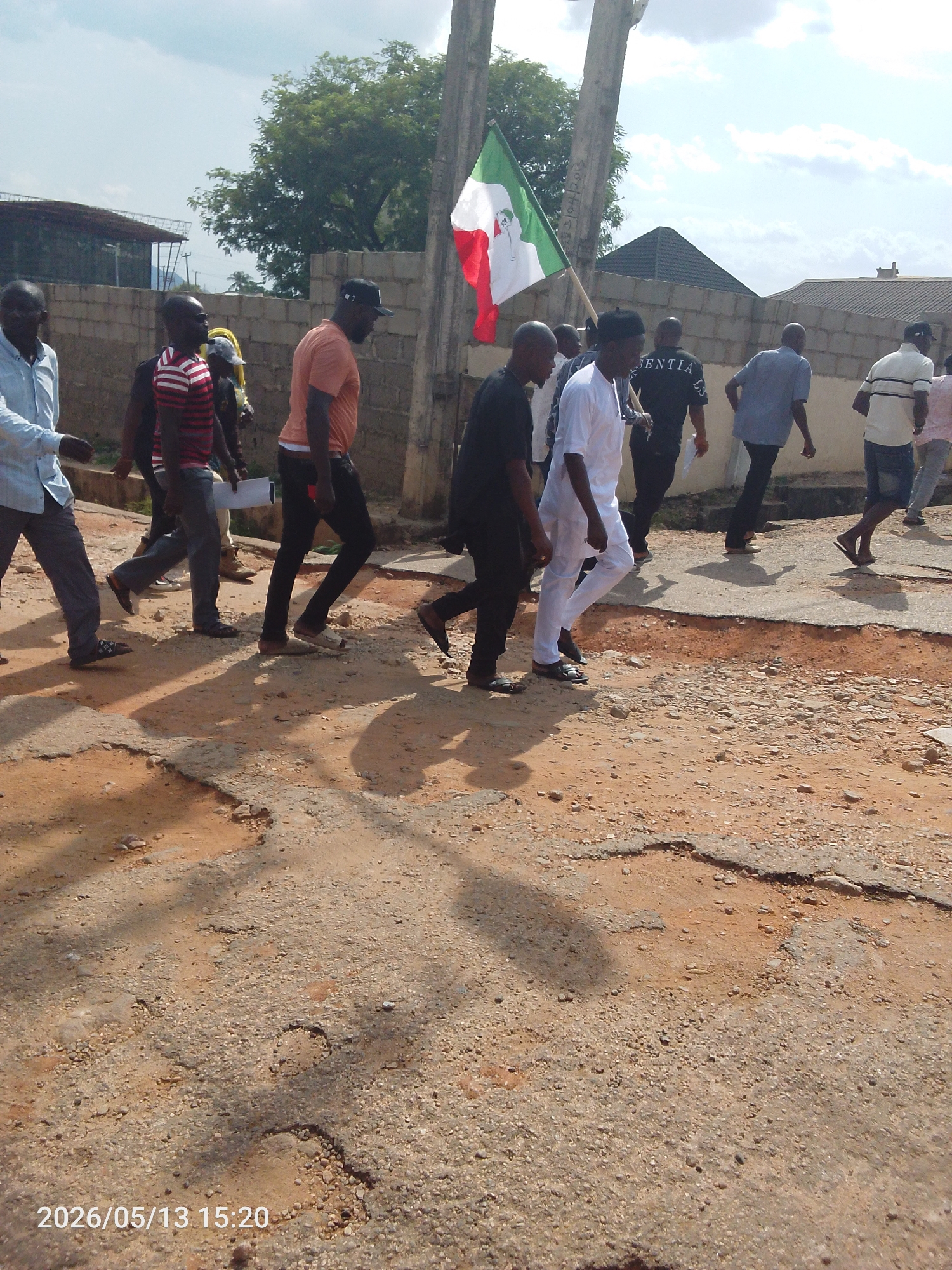 A group of about a dozen people are walking along a cracked and damaged asphalt road in Adankolo, Nigeria. The road is patched and has visible potholes with loose gravel and dirt. The individuals are dressed in a variety of casual clothing, including shirts, pants, and sandals, with some wearing caps. A person in the middle of the group holds a flag with green, white, and red stripes, with an emblem in the white band. Behind the group, there is a low brick wall, houses with corrugated metal roofs, and trees under a partly cloudy sky. The shadows of the group and nearby poles are cast across the road. The date and time stamp "2026/05/13 15:20" is at the bottom left corner of the image.