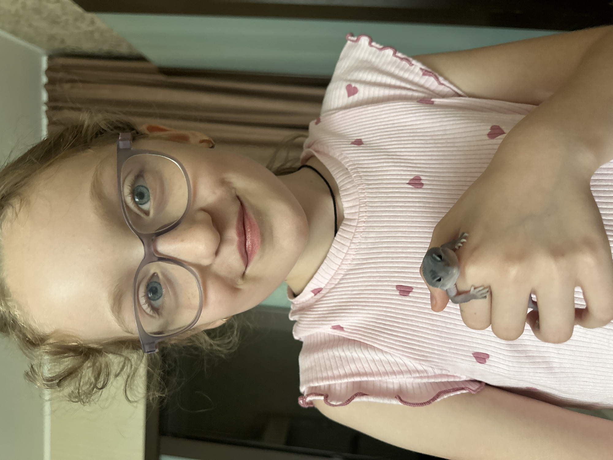 A young girl with blonde curly hair and blue eyes, wearing purple glasses and a pink ribbed t-shirt with small pink hearts, is holding a small grey lizard on her left hand. The girl is looking at the camera with a slight smile. The background appears to be an indoor setting with curtains and a wall. The image is a close-up shot, focusing on the girl and the lizard. There is no information available about the specific location within Shchyolkovo, Russia.