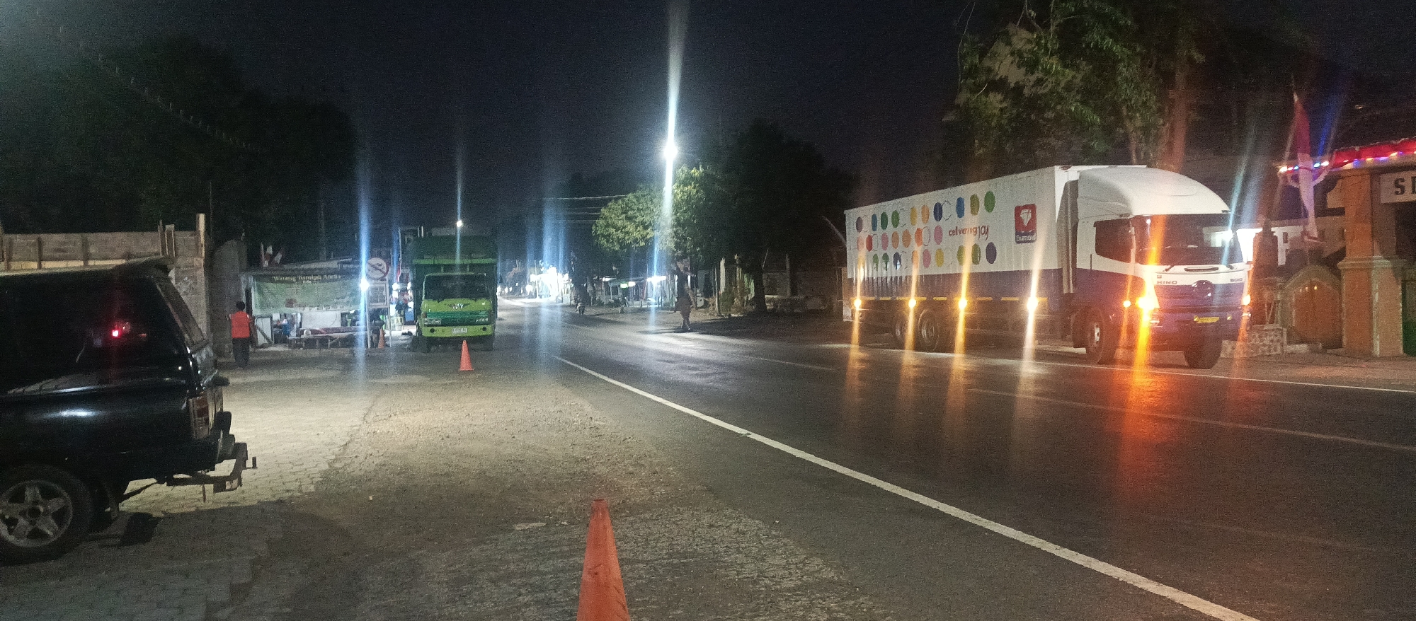 The image captures a nighttime scene on a street in Arjasa, Indonesia. A dark-colored SUV is parked on the left side of the road. Further down the street, a green truck is visible, with an orange traffic cone in front of it. To the right of the green truck, there is a large white and blue truck with a colorful pattern on its side, featuring the "Diamond" logo. The truck has its headlights and some side lights illuminated. A person in an orange vest is standing near a stall on the left side of the road. In the background, trees are present, and there are streetlights casting a glow on the asphalt road. A gate or entrance with a sign that reads "SI" is visible on the far right. The overall atmosphere suggests a street scene with ongoing traffic and some activity near a roadside establishment.