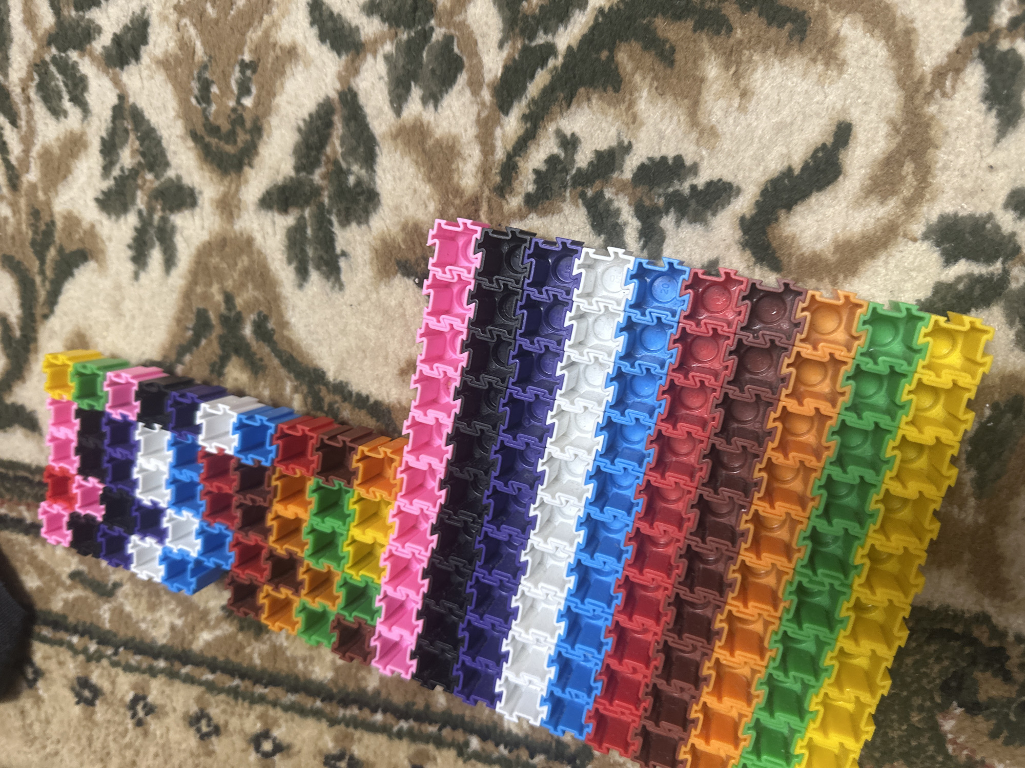 The image shows a close-up, overhead view of a collection of colorful, interlocking plastic building blocks arranged on a carpet. The blocks are stacked in rows, forming a structure that appears to be a ramp or a long, ascending path. The colors of the blocks include yellow, green, orange, brown, red, blue, white, purple, black, and pink, arranged in a distinct sequence. The carpet has a patterned design with brown and cream-colored swirls. The scene is indoors, and there are no people or animals visible. The lighting suggests it's daytime, possibly in a residential setting. No specific location in Загородный, Russia can be determined from the visual information provided.