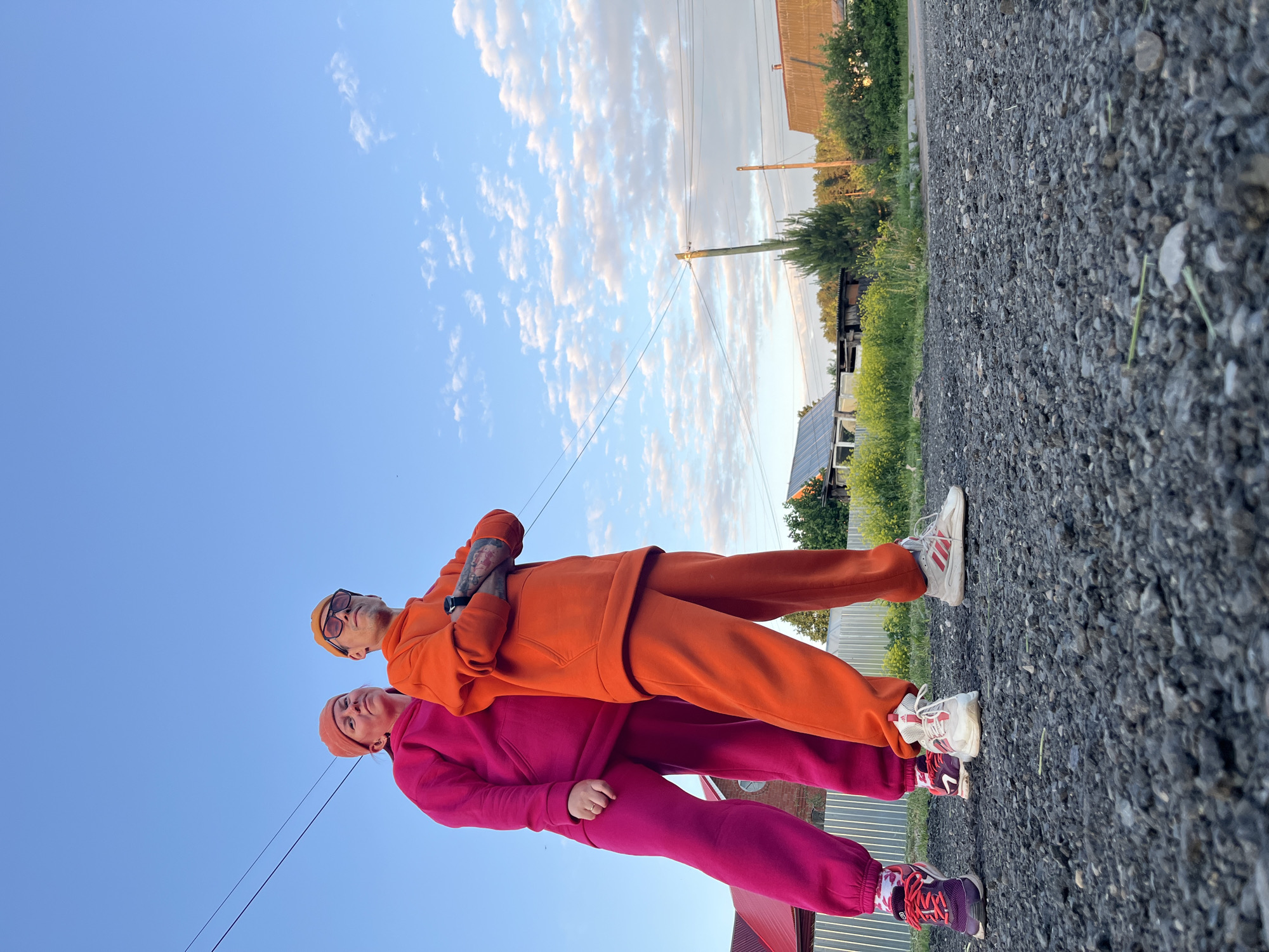 The image, rotated 90 degrees clockwise, captures two individuals standing on a gravel path outdoors, likely in Chaikovsky, Russia.
The foreground features two adults, both wearing brightly colored tracksuits with matching beanies. The person closer to the camera is dressed in a fuchsia or hot pink tracksuit and beanie, paired with multi-colored sneakers, pink laces, and patterned socks. The individual behind them wears an orange tracksuit and beanie, sunglasses, and white sneakers with red accents, resembling an Adidas logo. Tattoos are visible on the left forearm of the person in orange, who stands with arms crossed.
The gravel path extends into the background, flanked by green grass and yellow wildflowers. The surrounding environment includes lush green trees, various buildings with dark roofs, utility poles, and overhead power lines. A small array of solar panels is visible on one of the structures in the distance.
Above the scene, a vast expanse of clear blue sky is dotted with scattered fluffy white cumulus clouds, indicating fair weather. The warm, soft light suggests the time of day is late afternoon or early evening. The individuals appear to be casually posing for a photograph, conveying a friendly and relaxed atmosphere.