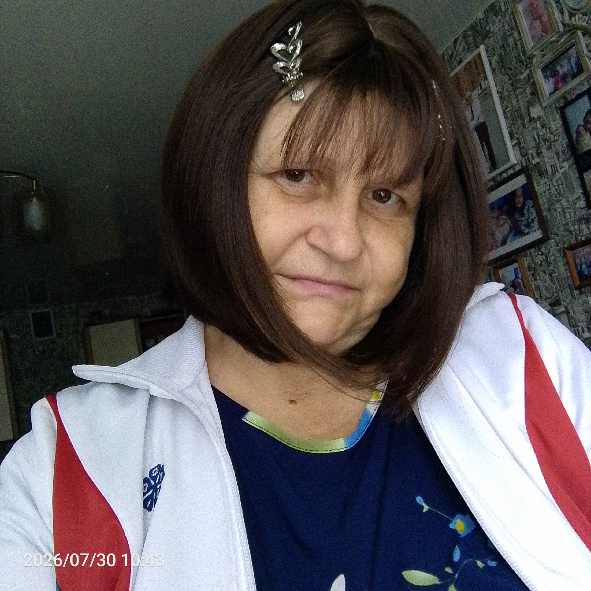 A middle-aged woman with dark brown hair is taking a selfie indoors. She is wearing a white and red athletic jacket over a dark blue shirt with a floral pattern. A silver hair clip with a heart design is in her hair. The background shows a wall with patterned wallpaper and framed pictures. A date stamp in the lower left corner reads "2026/07/30 10:43".