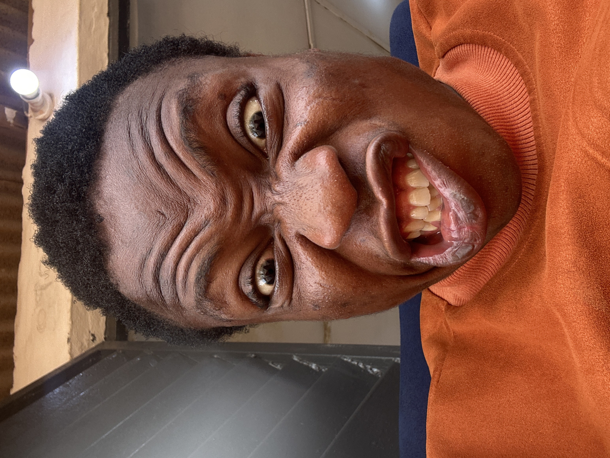 A close-up, rotated image captures a person's face with an intense expression. The individual has dark skin, closely cropped hair, and visible wrinkles on their forehead. Their eyes are wide and their mouth is open, revealing teeth. The person is wearing an orange top with a ribbed collar. The background includes a wall with a light fixture and a dark, possibly metallic, surface. The location and city are not discernible from the visual information.