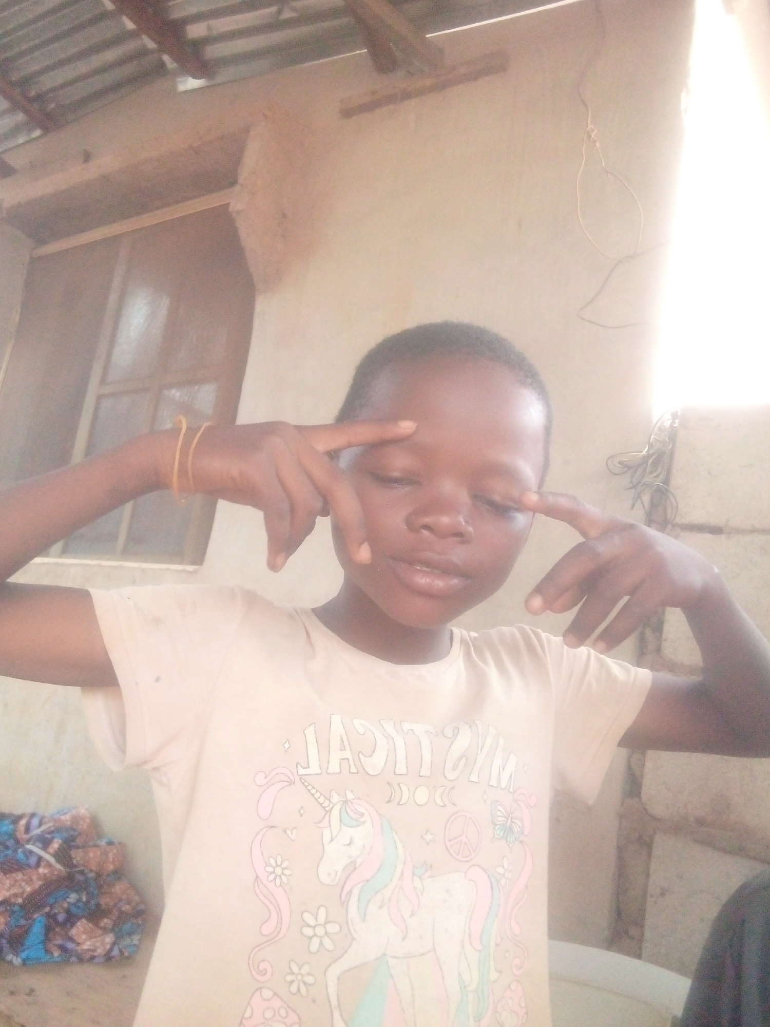 The image features a young child, likely a girl, posing confidently for the camera. She has dark skin, short dark hair, and is wearing a light-colored short-sleeved t-shirt. On her left wrist, she wears two thin yellow bands, possibly rubber bands. Her hands are raised with index fingers pointing towards her temples, a common playful or "thinking" gesture. Her eyes are slightly closed, and her lips are softly parted, conveying a relaxed and perhaps playful expression.
The t-shirt prominently displays a graphic design featuring a unicorn with a pink mane and blue tail, surrounded by various whimsical elements such as flowers, mushrooms, crescent moons, stars, a peace sign, and a butterfly. The text "MYSTICAL" is visible in a mirrored fashion across the chest, with "MAGICAL" potentially also present.
The background suggests a simple, semi-outdoor or indoor setting. To the left, there is a window with a wooden frame and what appears to be a screen or grille. Below the window, a stack of colorful patterned fabric, possibly traditional African print, is visible. Above, a corrugated metal roof supported by wooden beams can be seen. On the right side of the frame, there's a strong, blown-out light source, possibly sunlight entering through an opening, illuminating some exposed wires and a light-colored wall. A part of a white object, possibly a basin, is visible at the bottom right.
The bright lighting indicates it is daytime. The rustic elements in the background, combined with the child's attire and pose, create a candid and natural portrait.