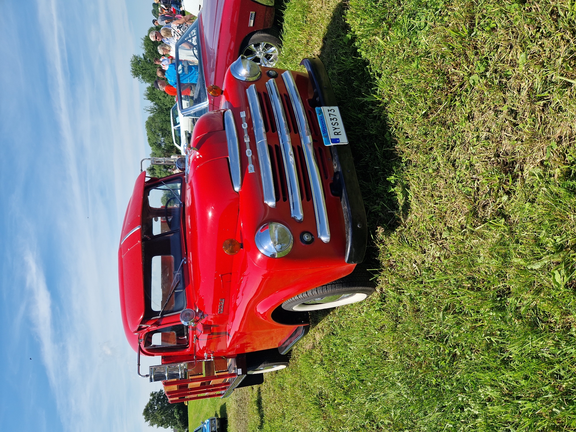 The image shows a bright red vintage Dodge truck parked on a grassy field under a partly cloudy blue sky. The truck has a wooden bed and chrome accents on the grille and headlights. A Minnesota license plate with the number RYS373 is visible on the front bumper. In the background, other vehicles and people are gathered, suggesting an outdoor event or car show. The location is identified as Edenberga, Sweden. The scene is well-lit by natural daylight.