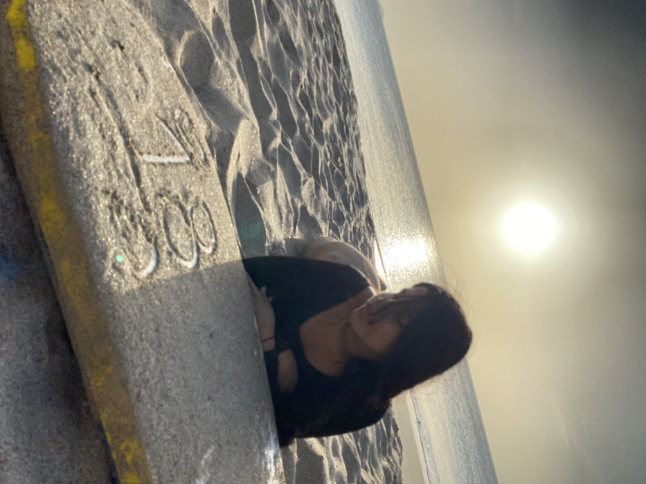 The image depicts a solitary female subject on a sandy beach during either sunrise or sunset in Juan Griego, Venezuela. The young female, with long, dark hair, is positioned prone on the light-colored sand, her body oriented towards the distant water. Her head is turned slightly to her right, and her gaze is directed downwards towards an object in the immediate foreground. She is dressed in a dark, long-sleeved top and light-colored bottoms. Her forearms rest on a rectangular object, possibly a boogie board, which is partially covered with sand. The numbers "183" are visibly etched into the sand on the surface of this object. In the background, the sea stretches under a bright, low-hanging sun, which creates a prominent reflection path on the water's surface. The sky transitions from a brilliant yellow-orange near the horizon to a softer, muted gray higher up.